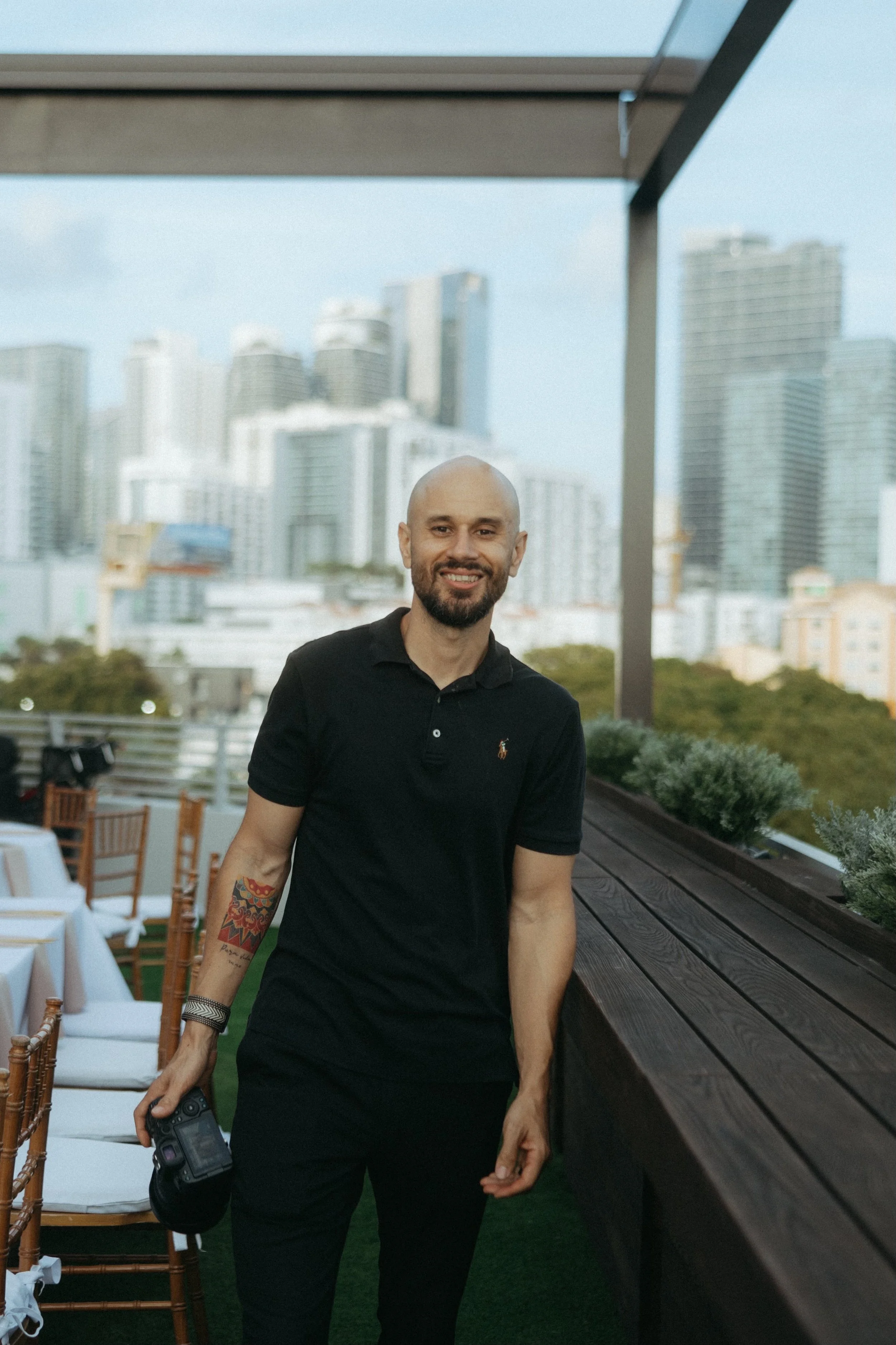 Man in black polo shirt holding a camera, standing on a rooftop with a cityscape skyline in the background.