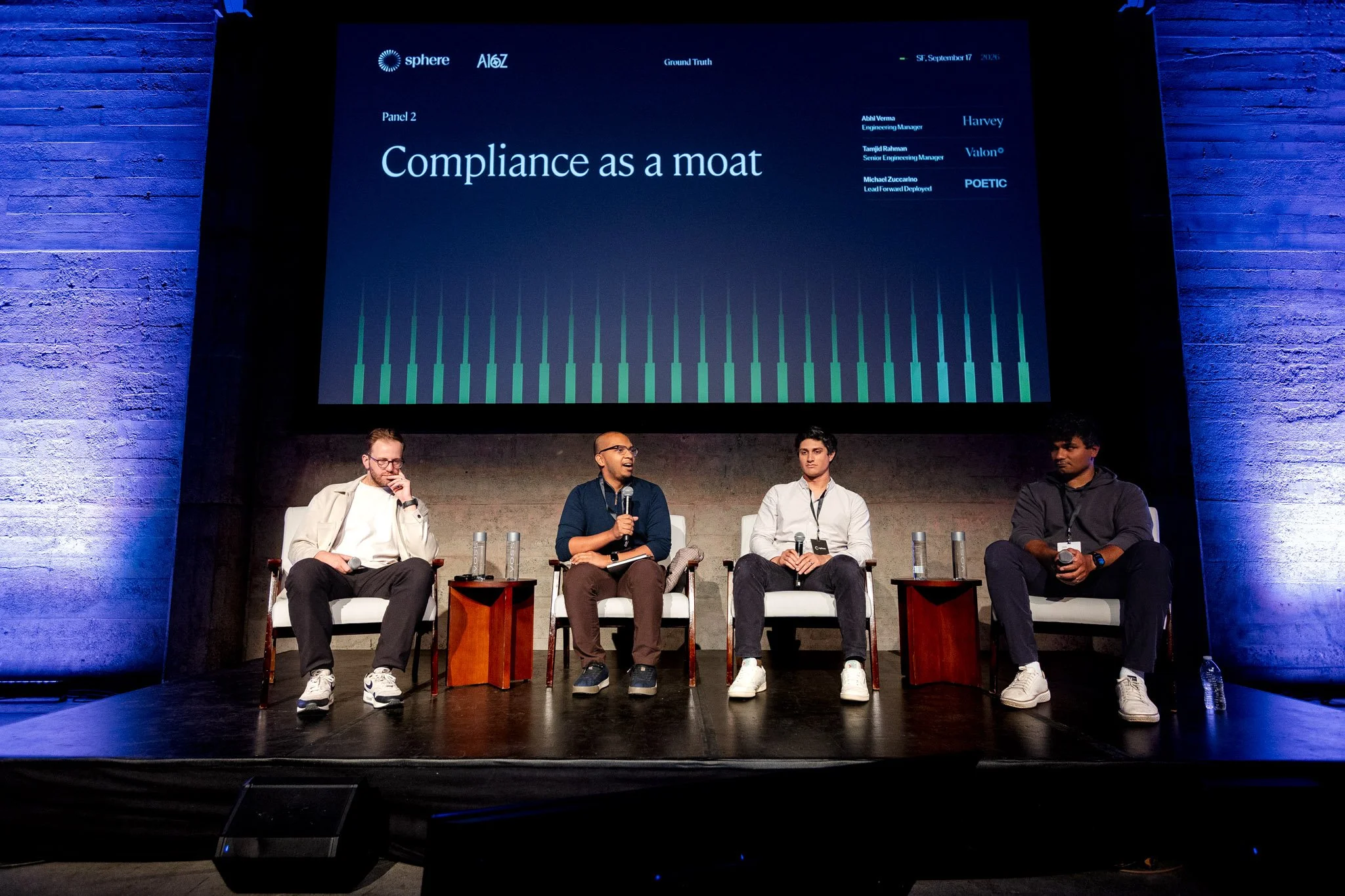 Four men sitting on chairs on a stage during a panel discussion with a screen behind them. The screen displays logos and the phrase "Compliance as a moat," along with panel information.