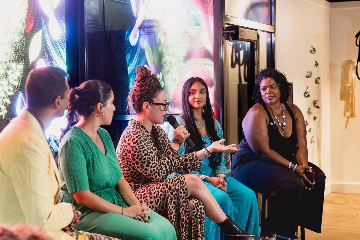 A group of six diverse women sitting in a semi-circle, engaged in a discussion or panel event, with one woman holding a microphone, on a stage with a colorful digital background.