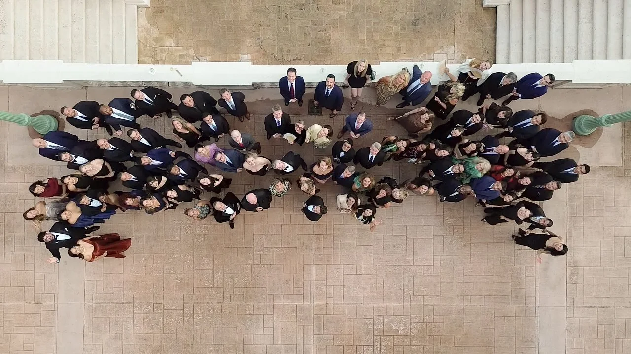 Group of hundreds of diverse people in formal attire standing outdoors in a formation that resembles an airplane, taken from above.