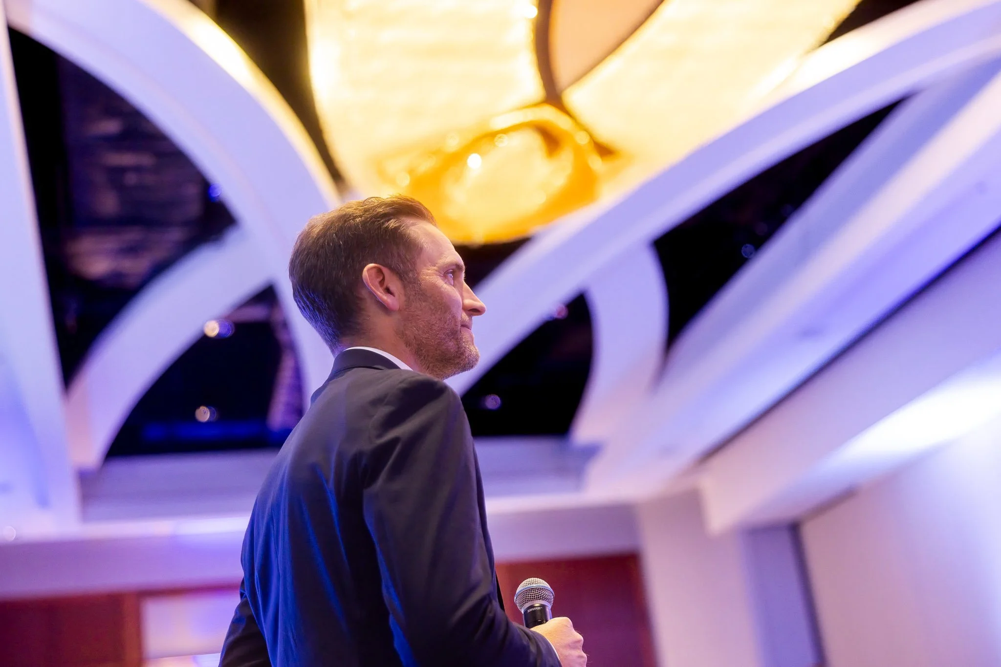 A man in a suit holding a microphone, standing in front of a decorative, abstract ceiling with warm and cool lighting.