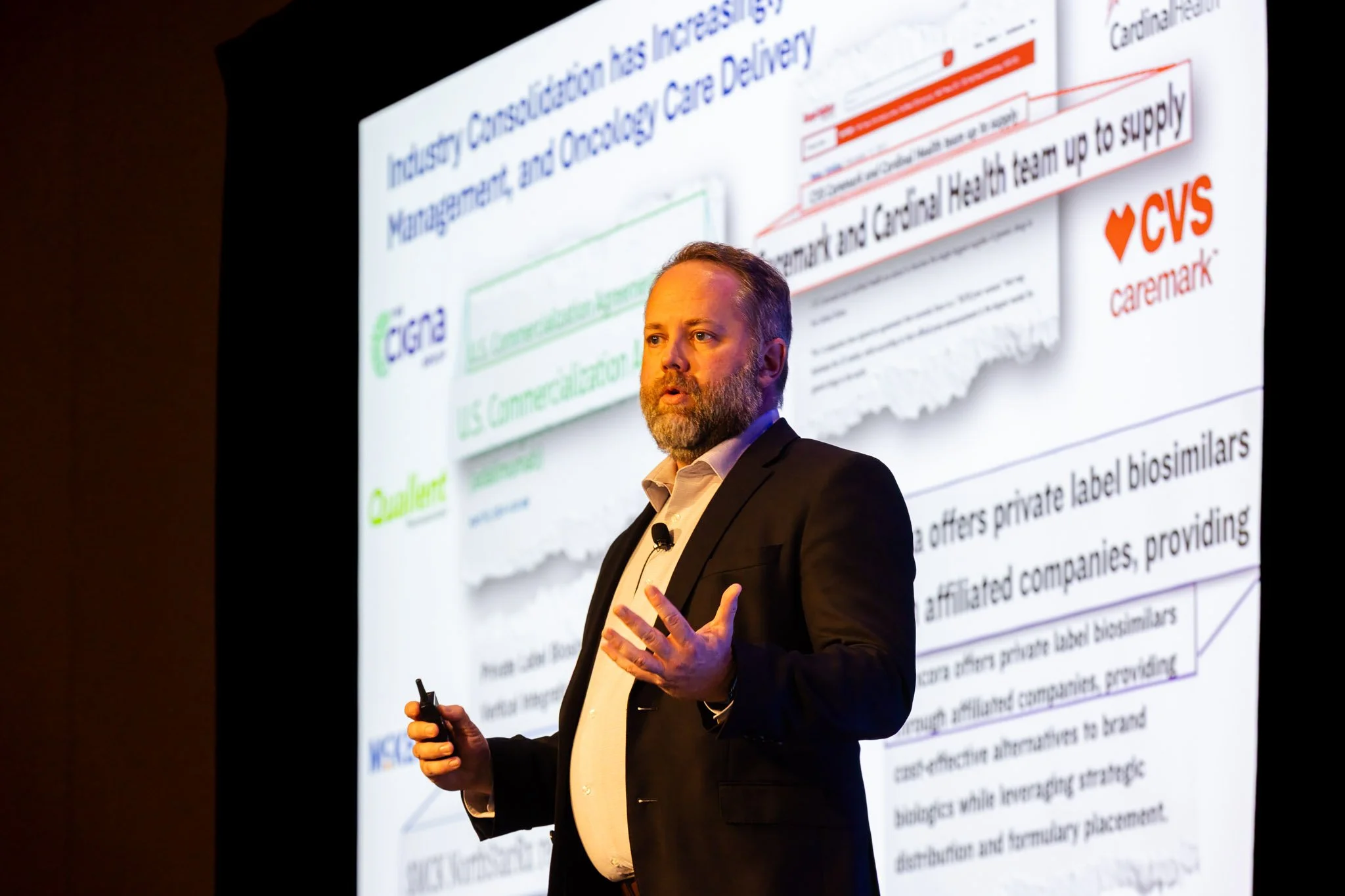 A man in a black suit giving a presentation in front of a large screen displaying a slide with text and logos, including CVS Caremark and other corporate branding.