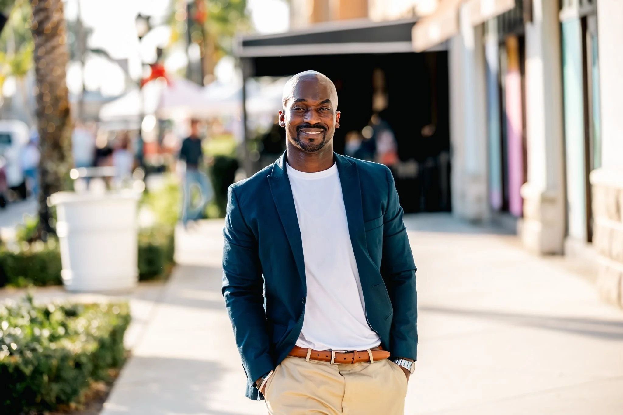 A man wearing a blue blazer, white T-shirt, tan pants, and a brown belt, smiling and walking outdoors on a sunny day with shops and people in the background.