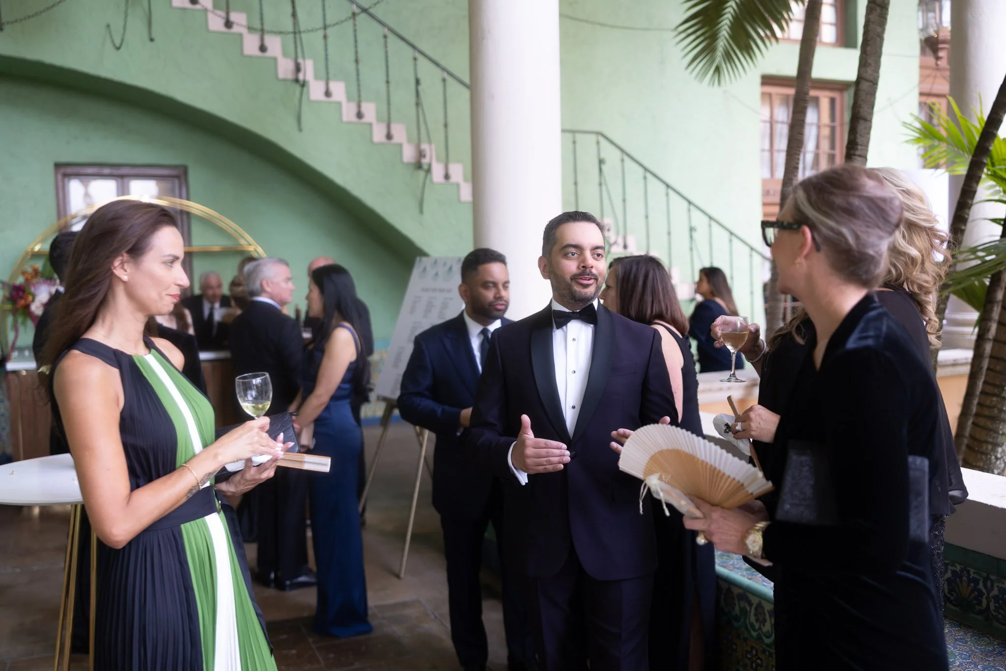 People dressed in formal attire socializing at a party, with some holding glasses of wine or fans, in an indoor space with green walls, staircase, and plants.