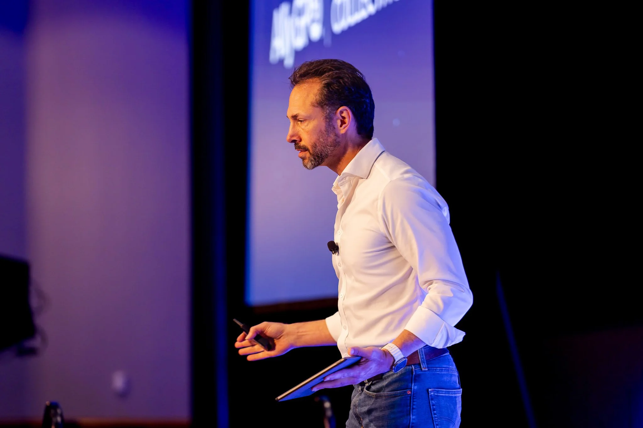 A man with dark hair and a beard wearing a white shirt and blue jeans, standing on stage during a presentation or speech, holding a tablet in his left hand and a remote control in his right. The background features a screen with partially visible tex