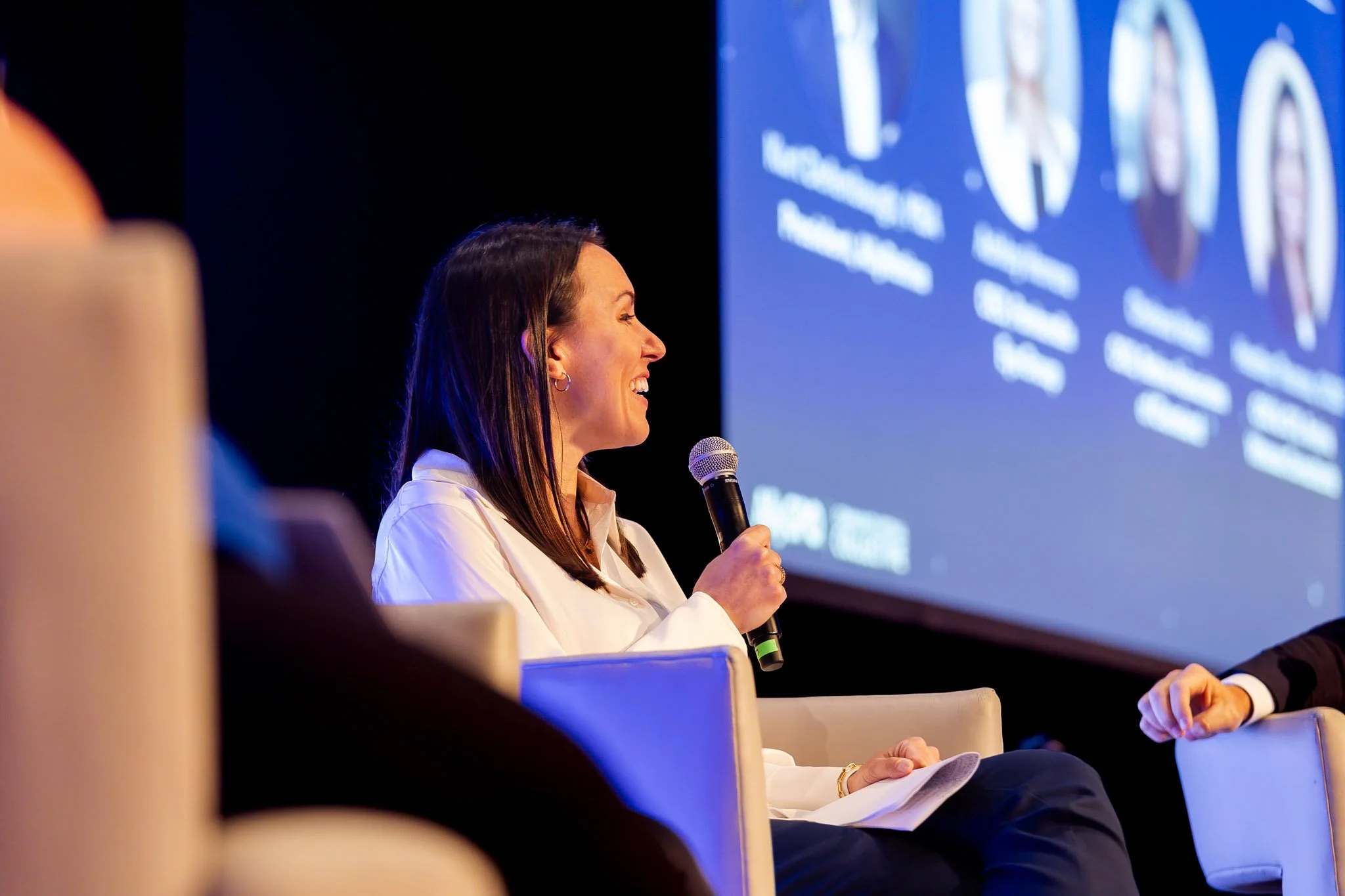 Side profile of a woman speaking into a microphone during a conference, with a large screen showing text and visuals in the background.