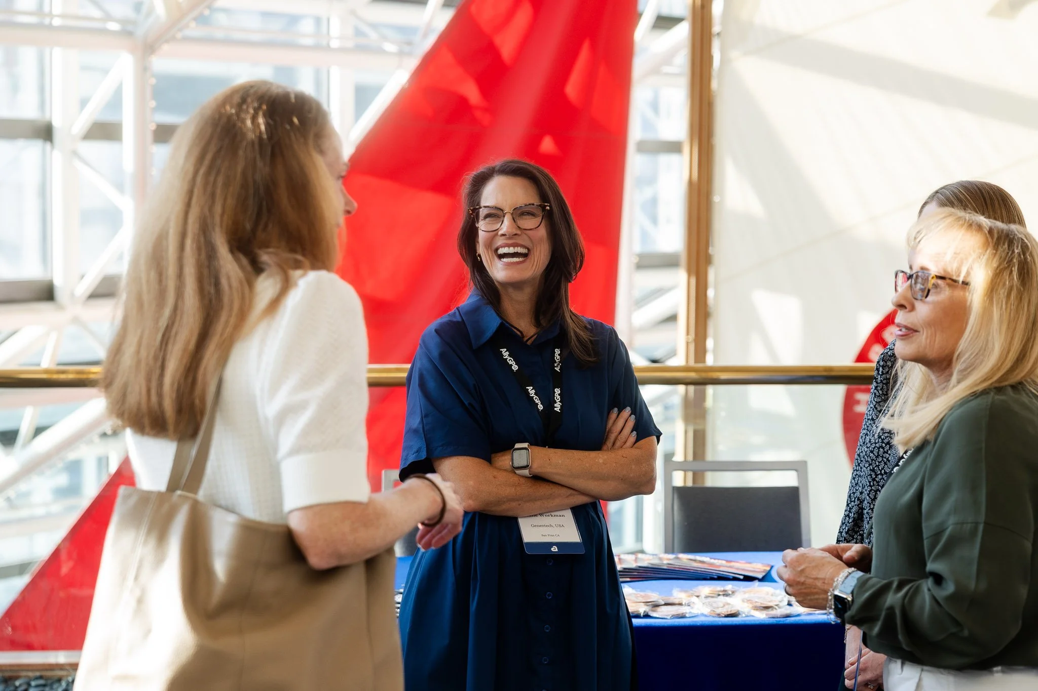Four women engaged in conversation at a conference or event, with tables and conference materials in the background, and natural light illuminating the space.