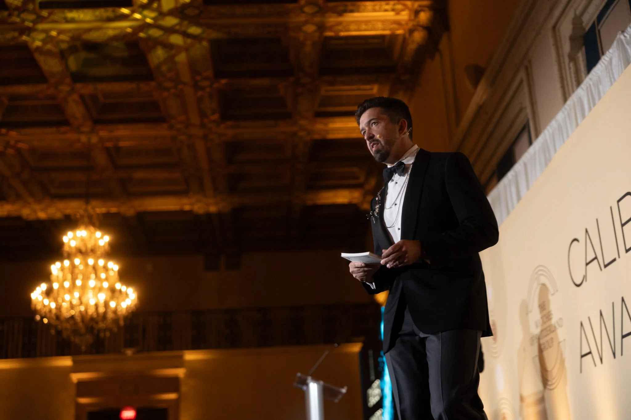 A man in formal attire, wearing a black tuxedo and bow tie, stands on stage at a formal event, holding a microphone and papers. The room has a high, ornate wooden ceiling and chandeliers hanging from above.