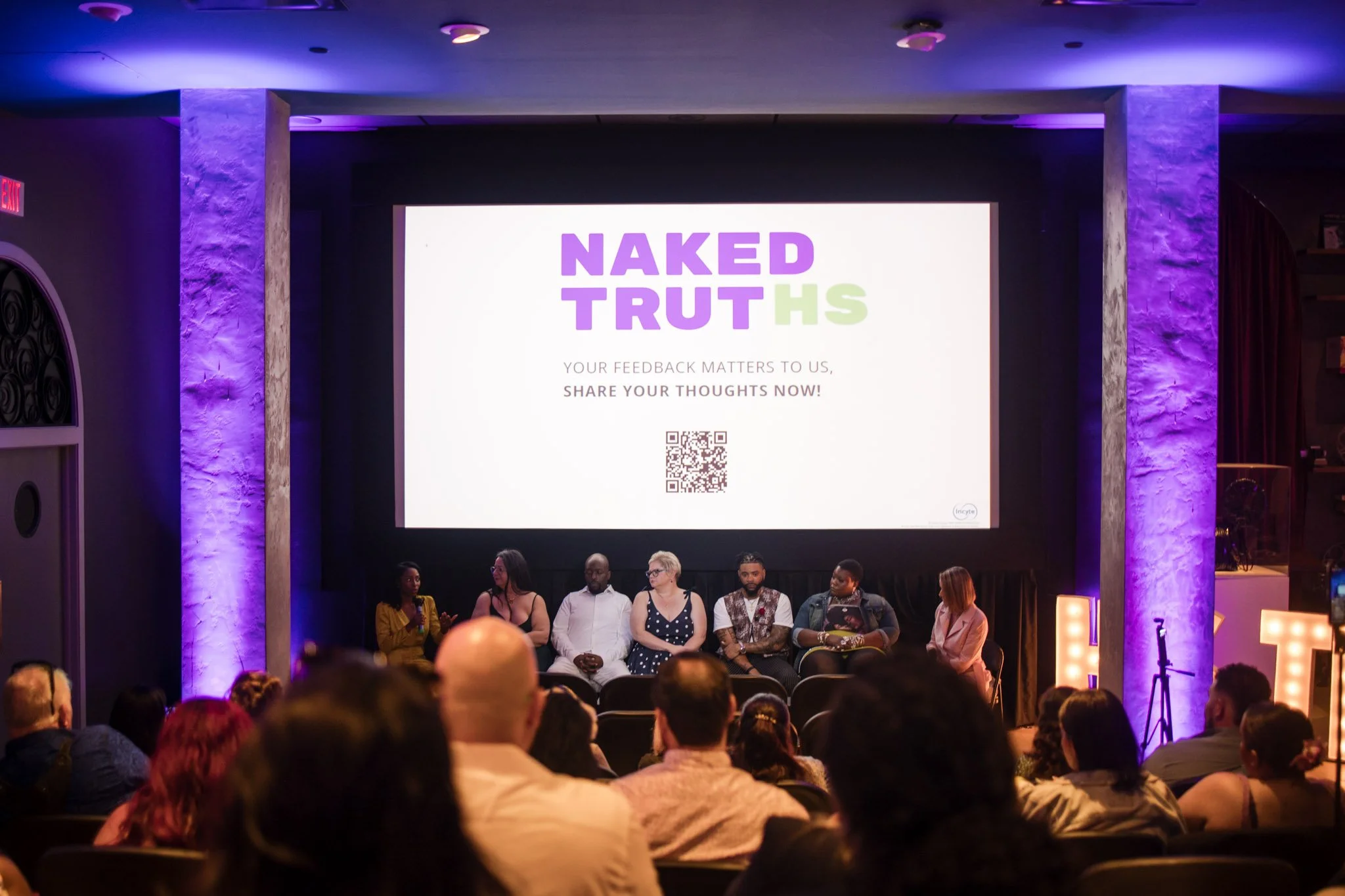 A group of people on stage participating in a panel discussion at an event called 'Naked Truths,' with a large screen behind them displaying the event name and a QR code. The audience is seated and listening attentively.