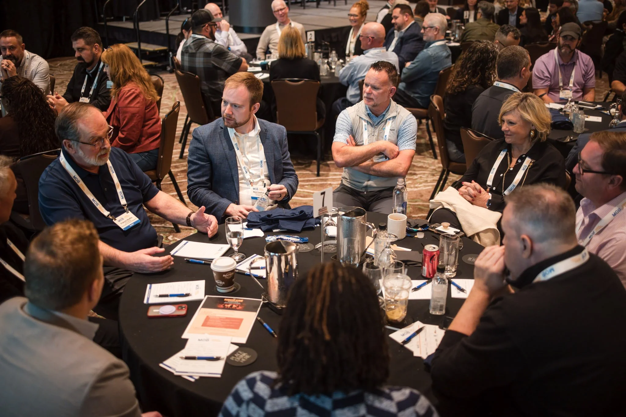 People seated around a round table at a conference or business event, engaging in discussion, with notepads, pens, and drinks on the table.