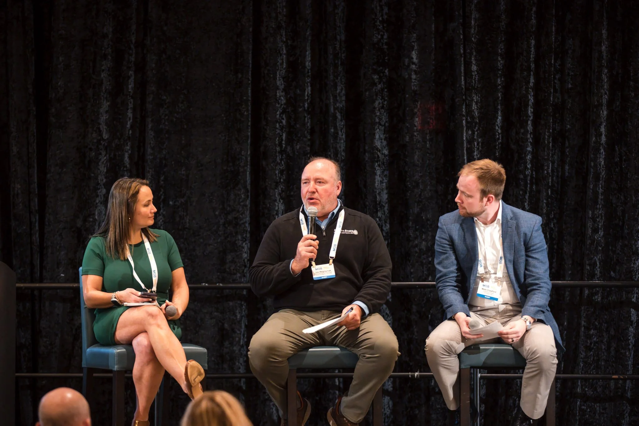Four people sitting on a stage in front of a black curtain at a panel discussion, with a man speaking into a microphone while the others listen.