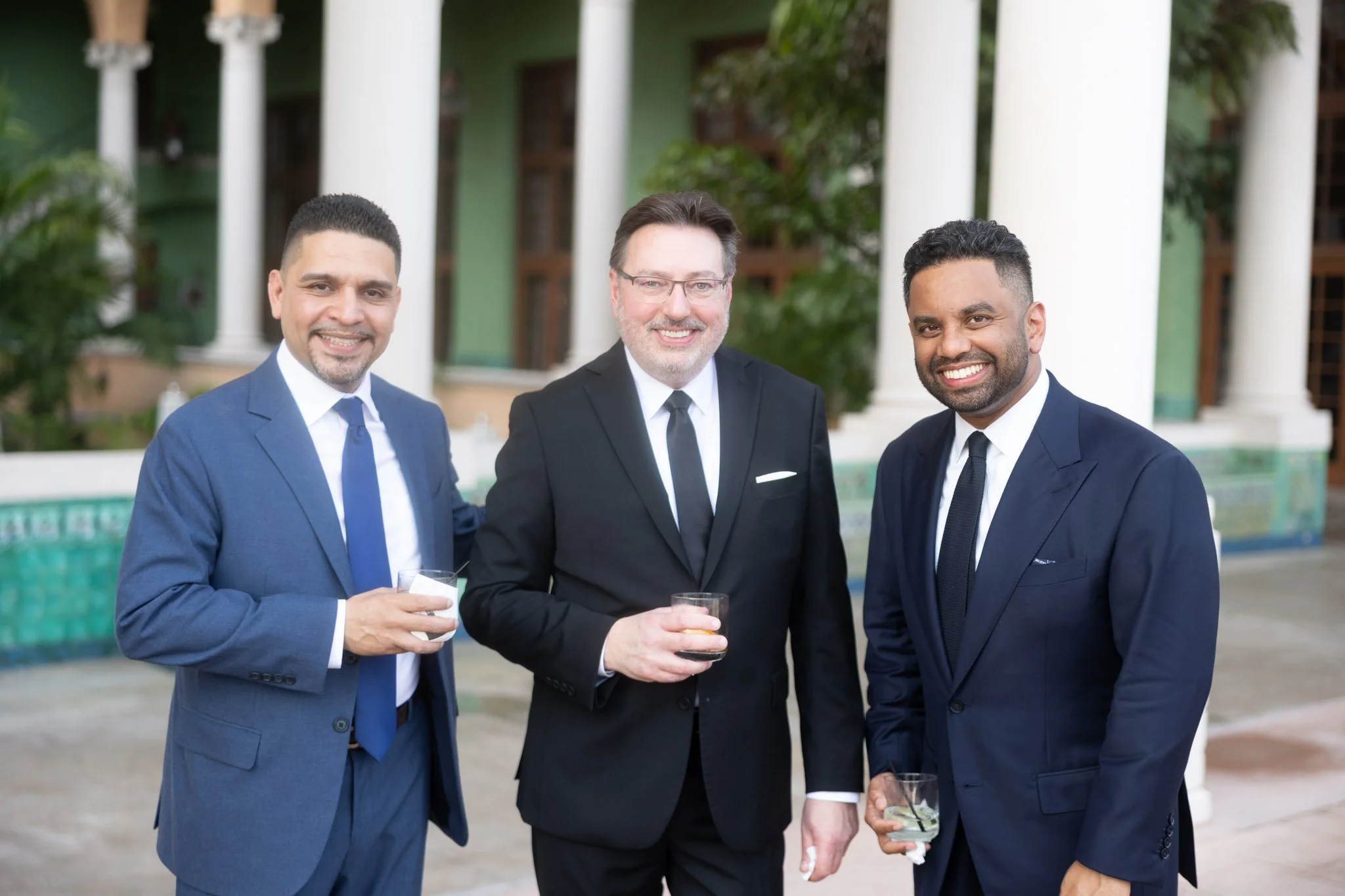 Three men in suits smiling and holding drinks at an outdoor event with a pool and columns in the background.