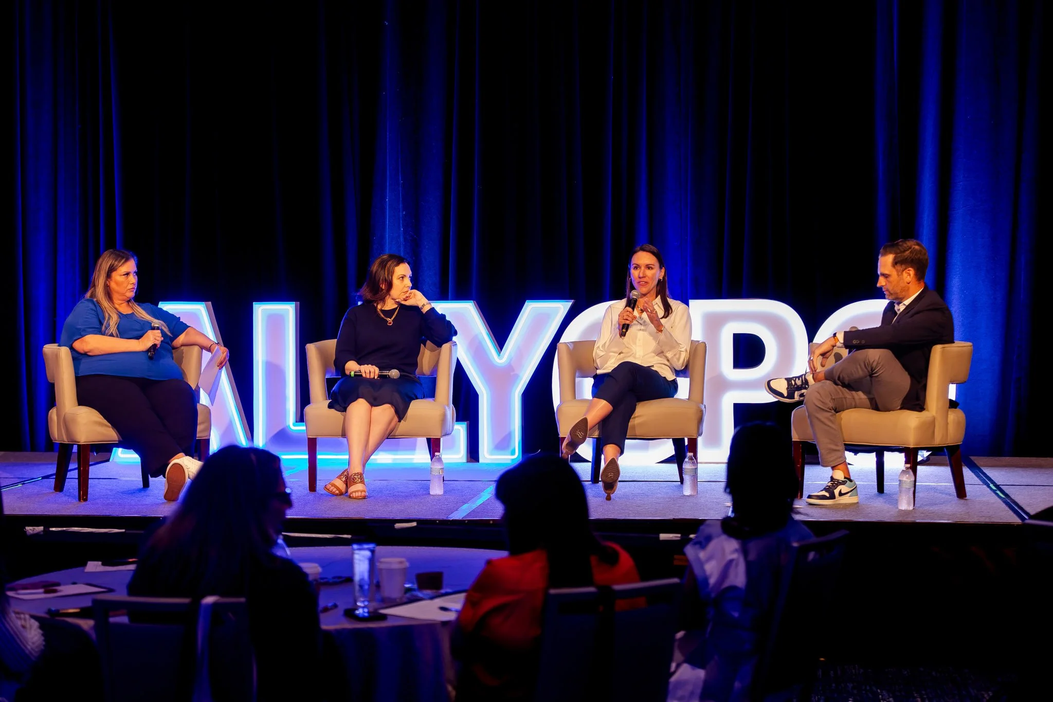 Four people sitting on chairs on stage engaging in a panel discussion at a conference, with a large illuminated sign behind them.