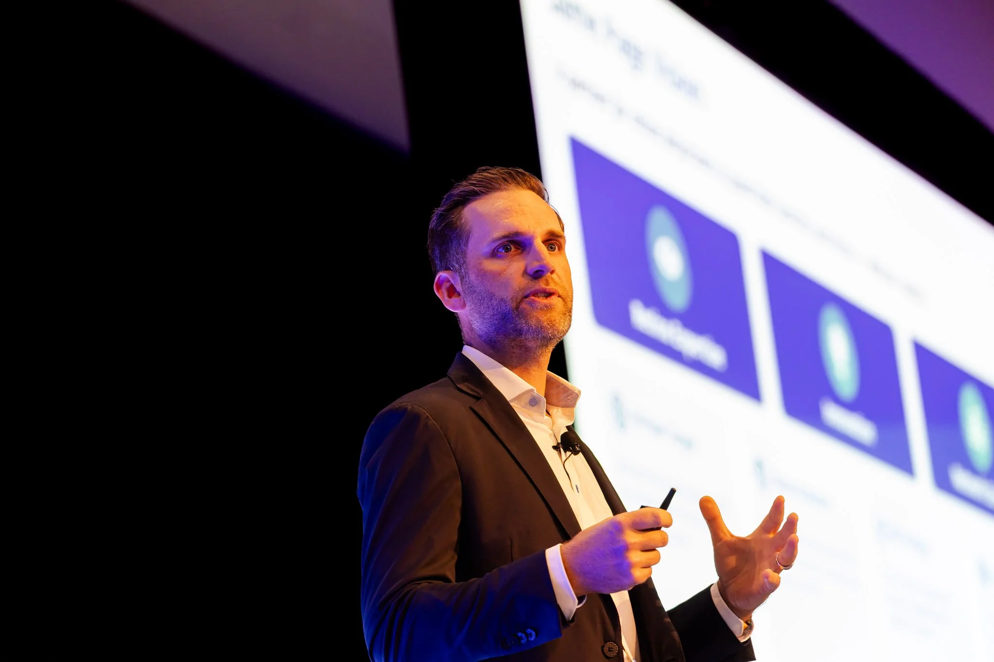 A man in a suit giving a presentation with a large screen behind him displaying slides.