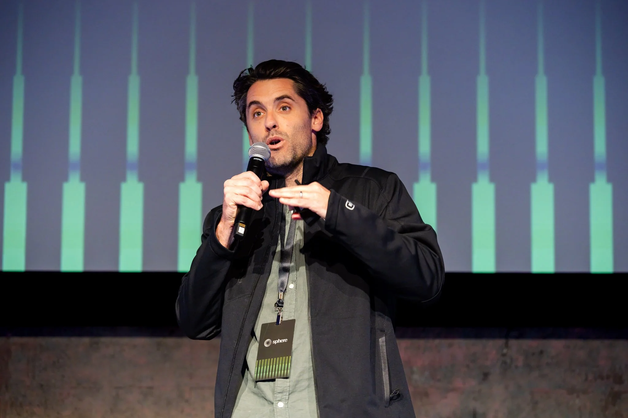 A man speaking into a microphone on stage, wearing a black jacket and a gray shirt with a conference badge around his neck. Background displays green vertical lines.