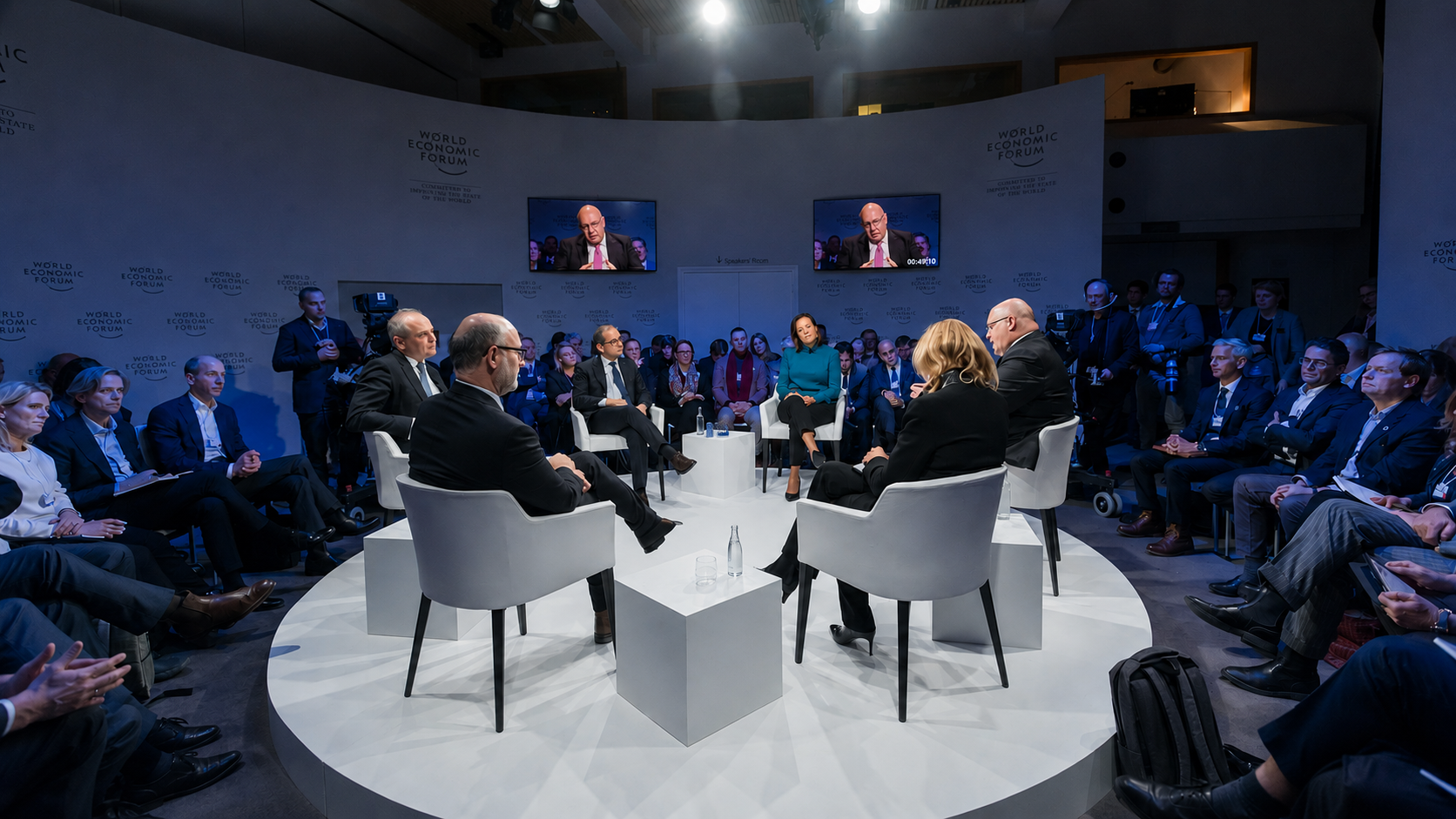A panel discussion at the World Economic Forum, with six panelists seated in white armchairs around a circular stage, an audience seated around the stage, and two large screens displaying a speaker.