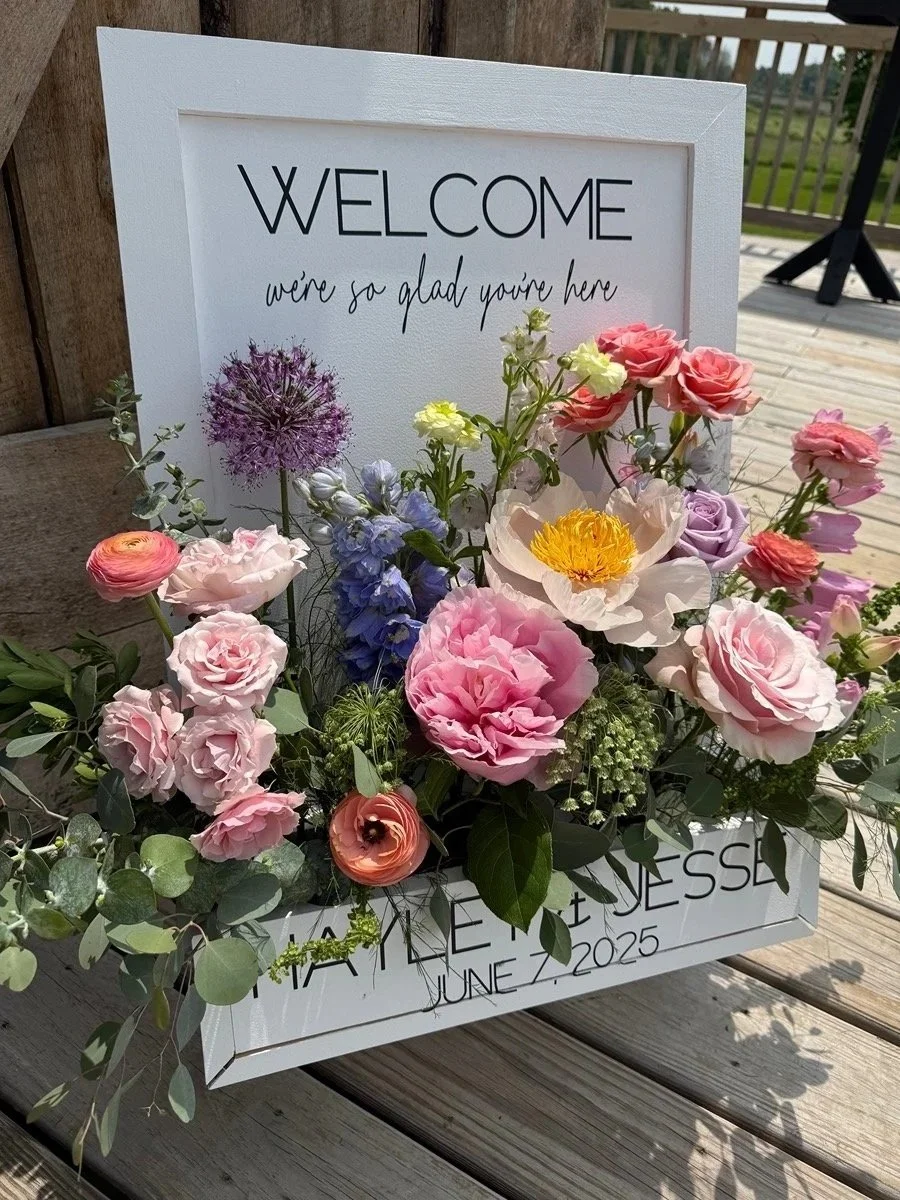 A white welcome sign with the message 'Welcome, we're so glad you're here' and a floral arrangement of pink, purple, yellow, and blue flowers on wooden deck.
