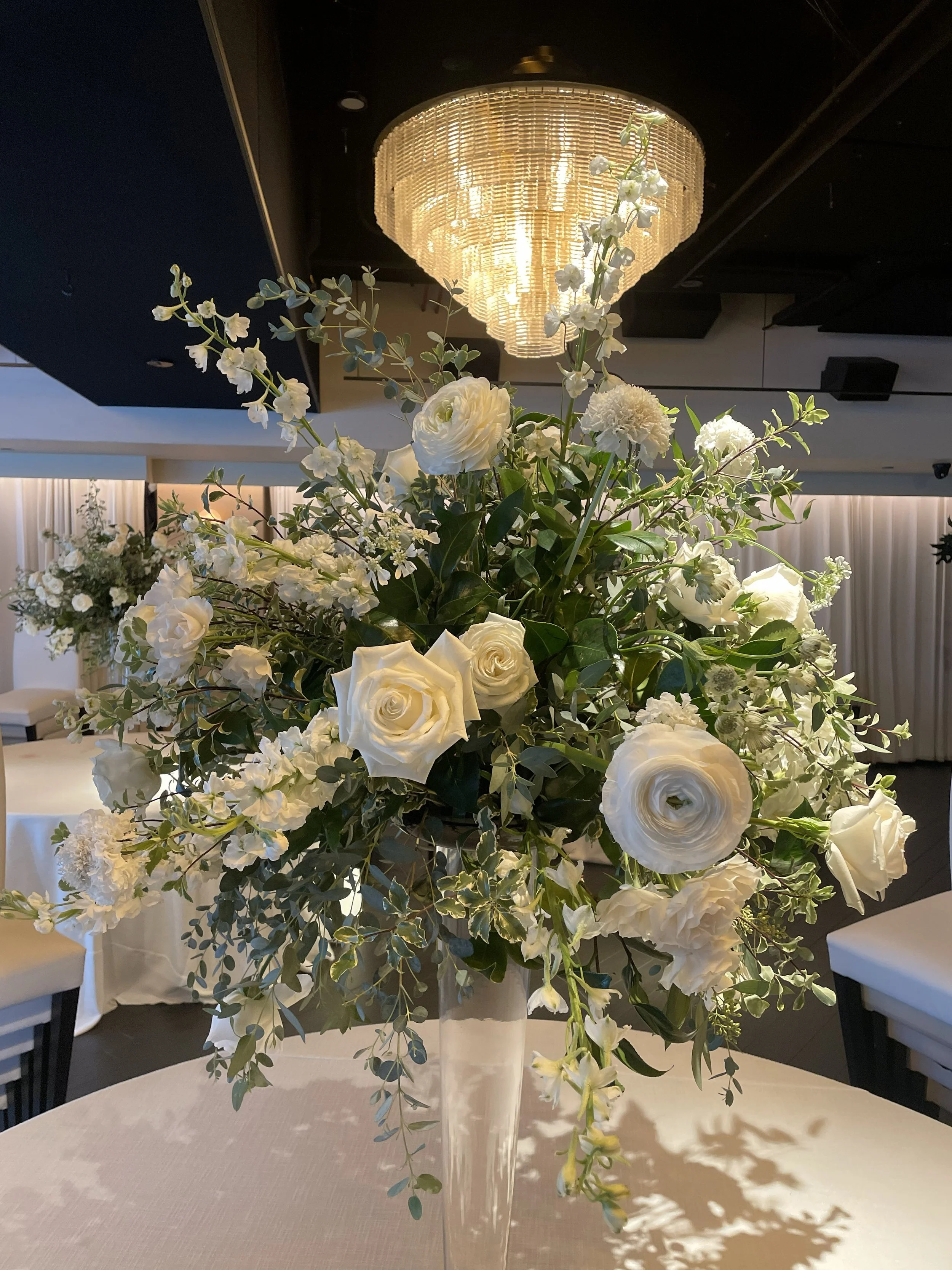 Elegant floral centerpiece with white roses, peonies, and greenery on a dining table, with a gold chandelier overhead.