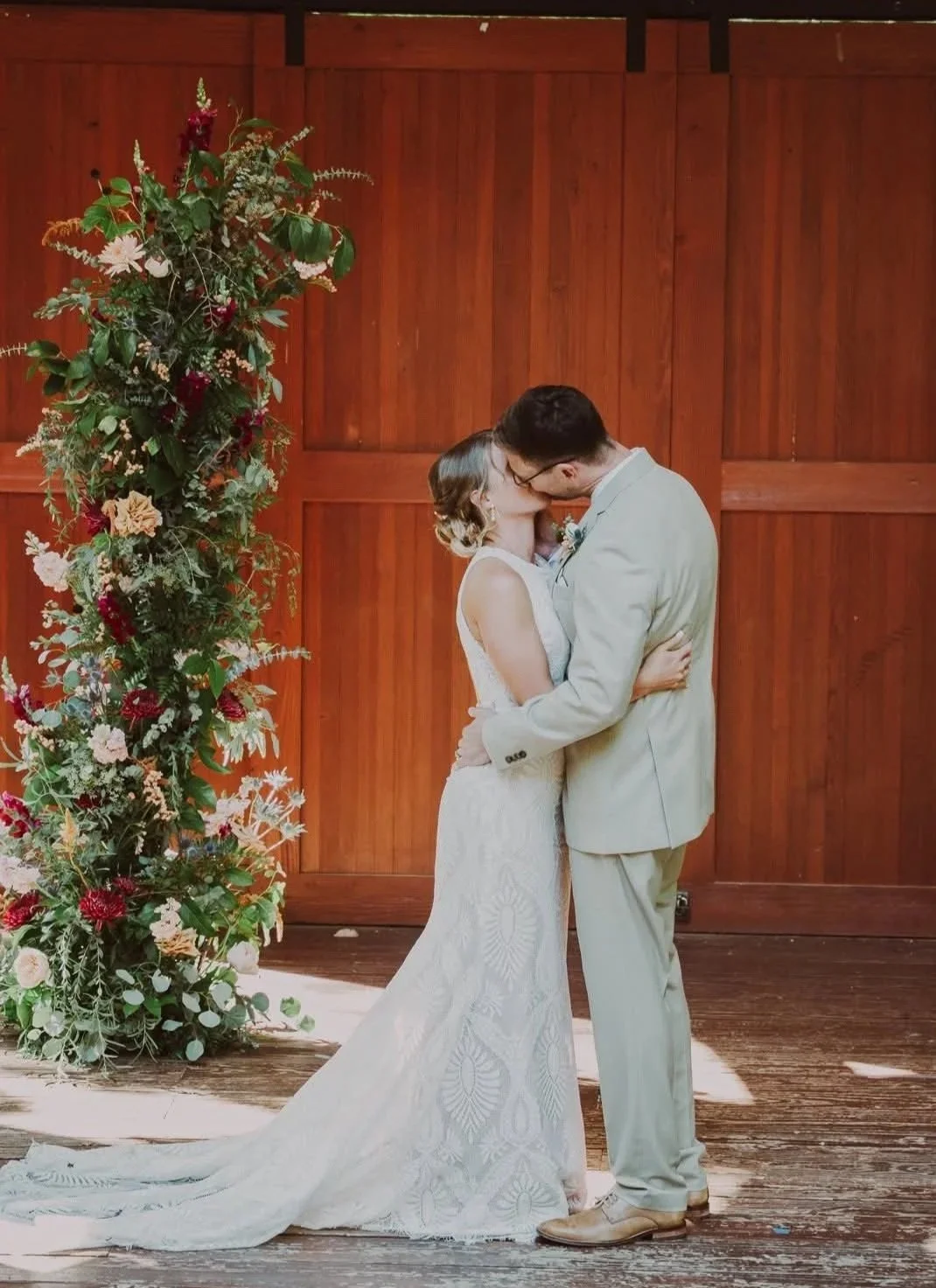 A couple kissing during their wedding ceremony in front of a wooden backdrop, with a floral arrangement on the side.