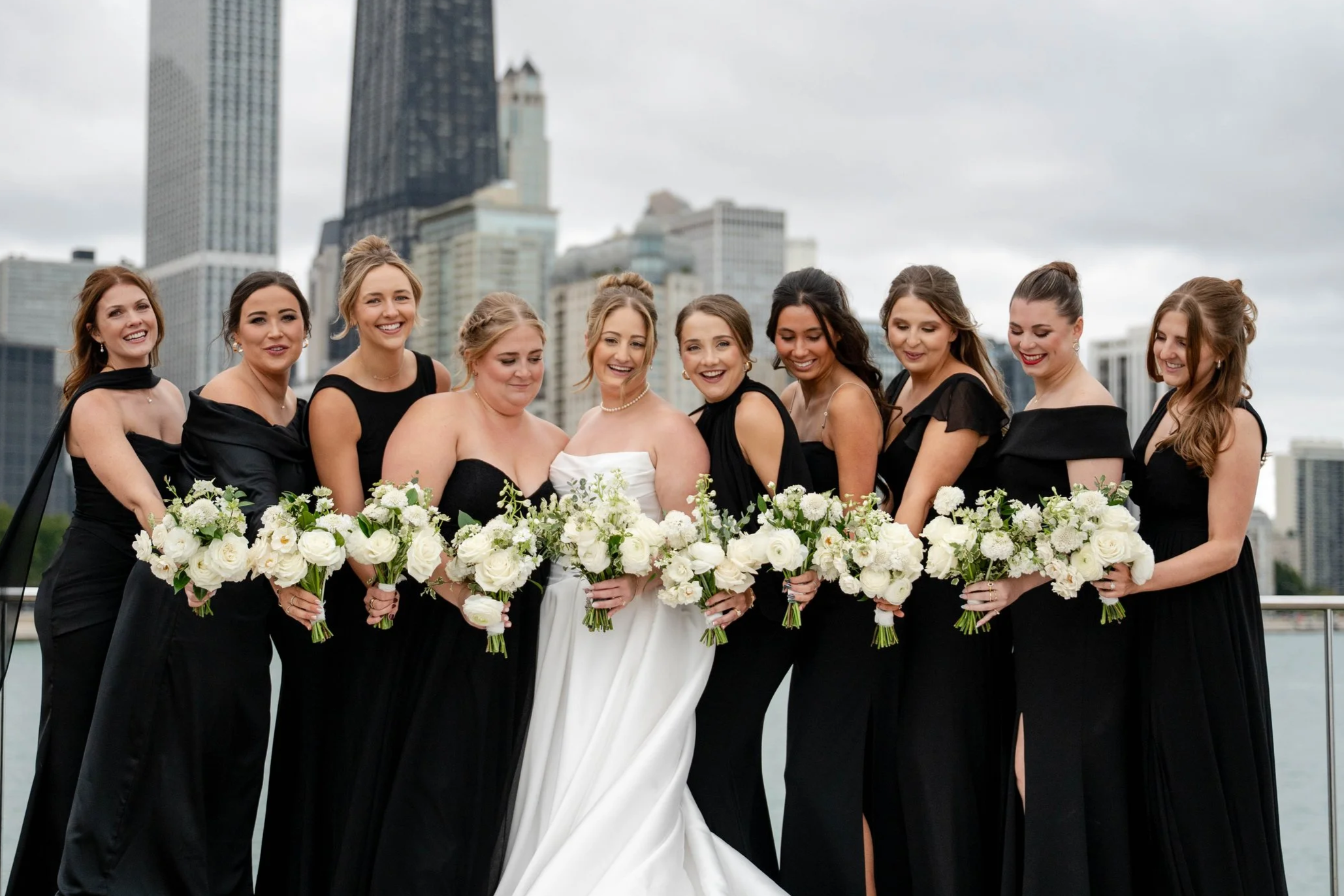 Bride in white wedding gown surrounded by friends in black dresses, holding white floral bouquets, with city skyline background.