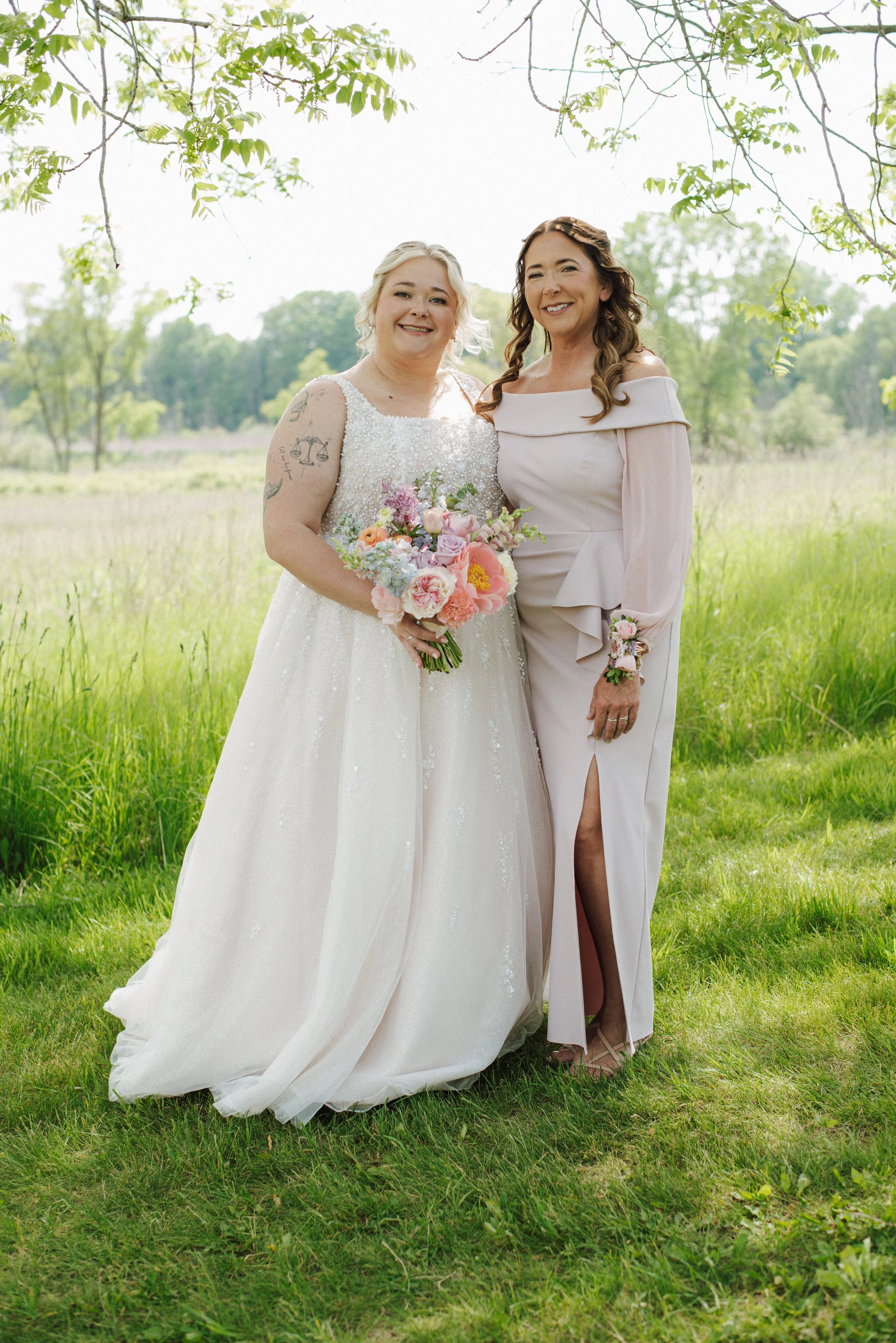 Two women standing outdoors in a grassy field, one in a wedding dress holding a bouquet, and the other in a pink off-shoulder dress with a slit, both smiling.
