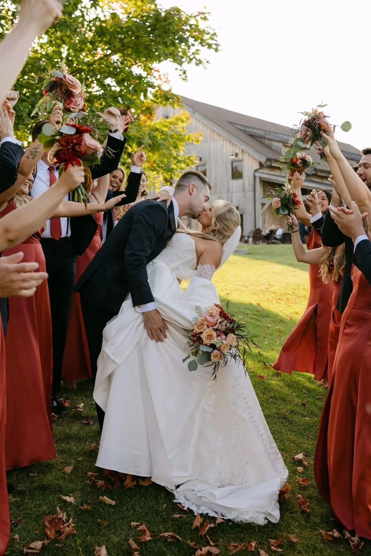 A bride and groom kissing under a shower of flowers during their wedding ceremony outdoors on a grassy area, surrounded by bridesmaids and groomsmen holding floral bouquets.