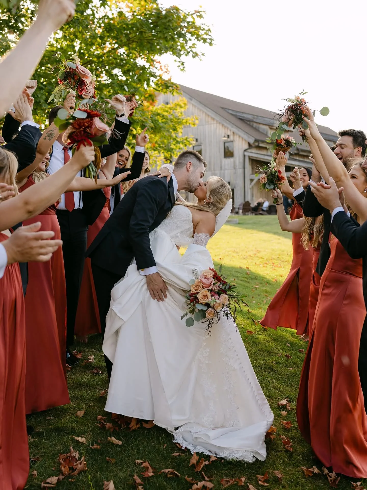 A newlywed couple kisses under a shower of bouquets held by friends and family in an outdoor wedding celebration.