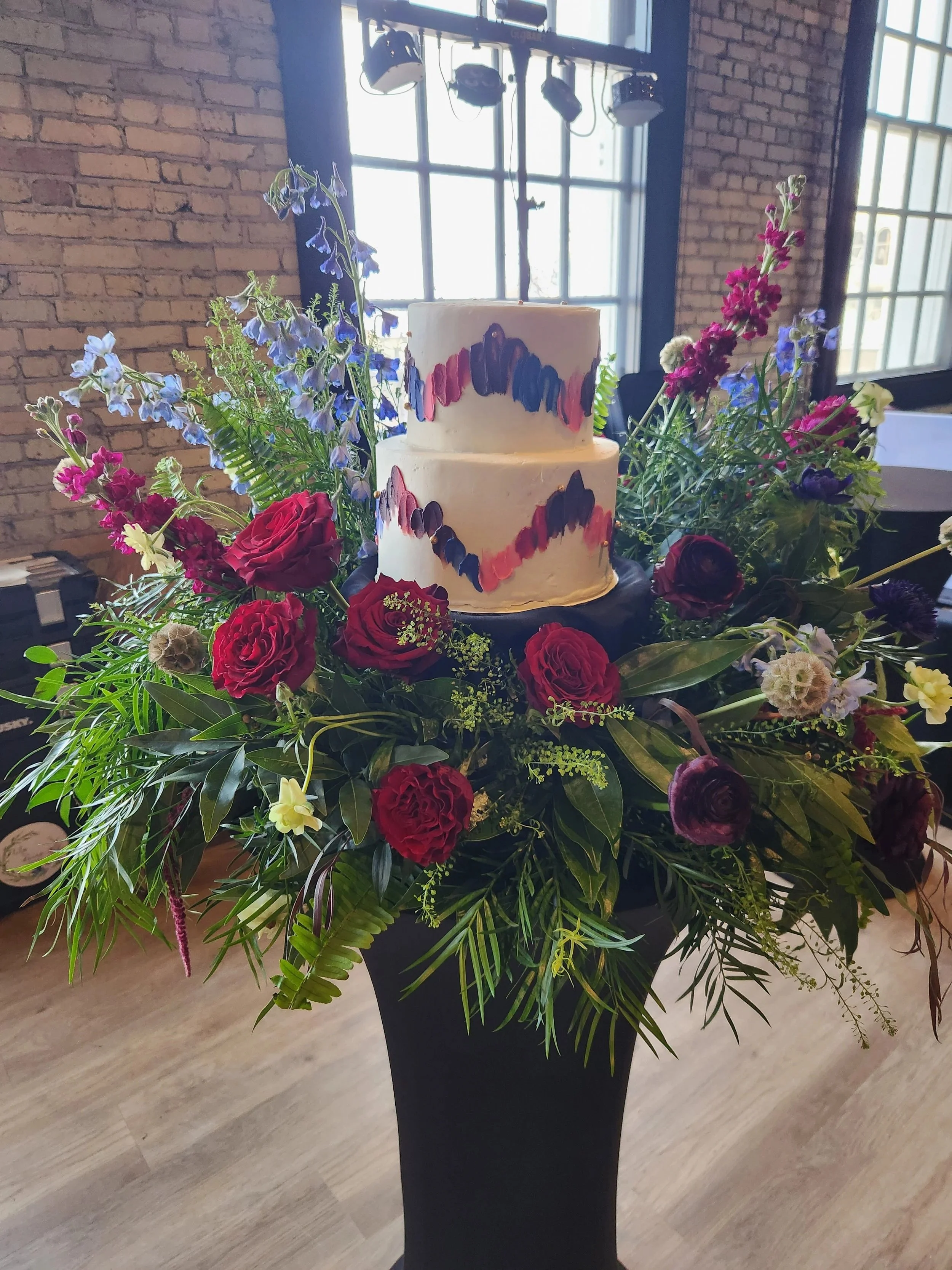 A two-tiered white cake with colorful brushstroke designs, surrounded by a large floral arrangement with red roses, purple and pink flowers, and green foliage, set in a black vase in front of brick walls and large windows.
