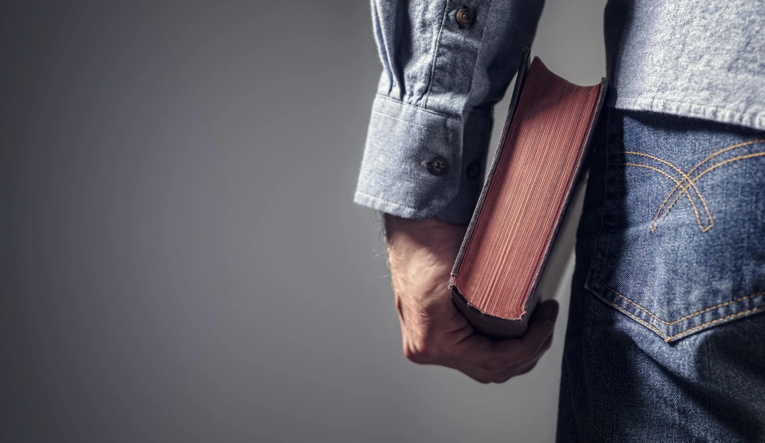 A person wearing a denim jacket with rolled-up sleeves and blue jeans, holding a closed hardcover book with a red spine and textured cover in their right hand.