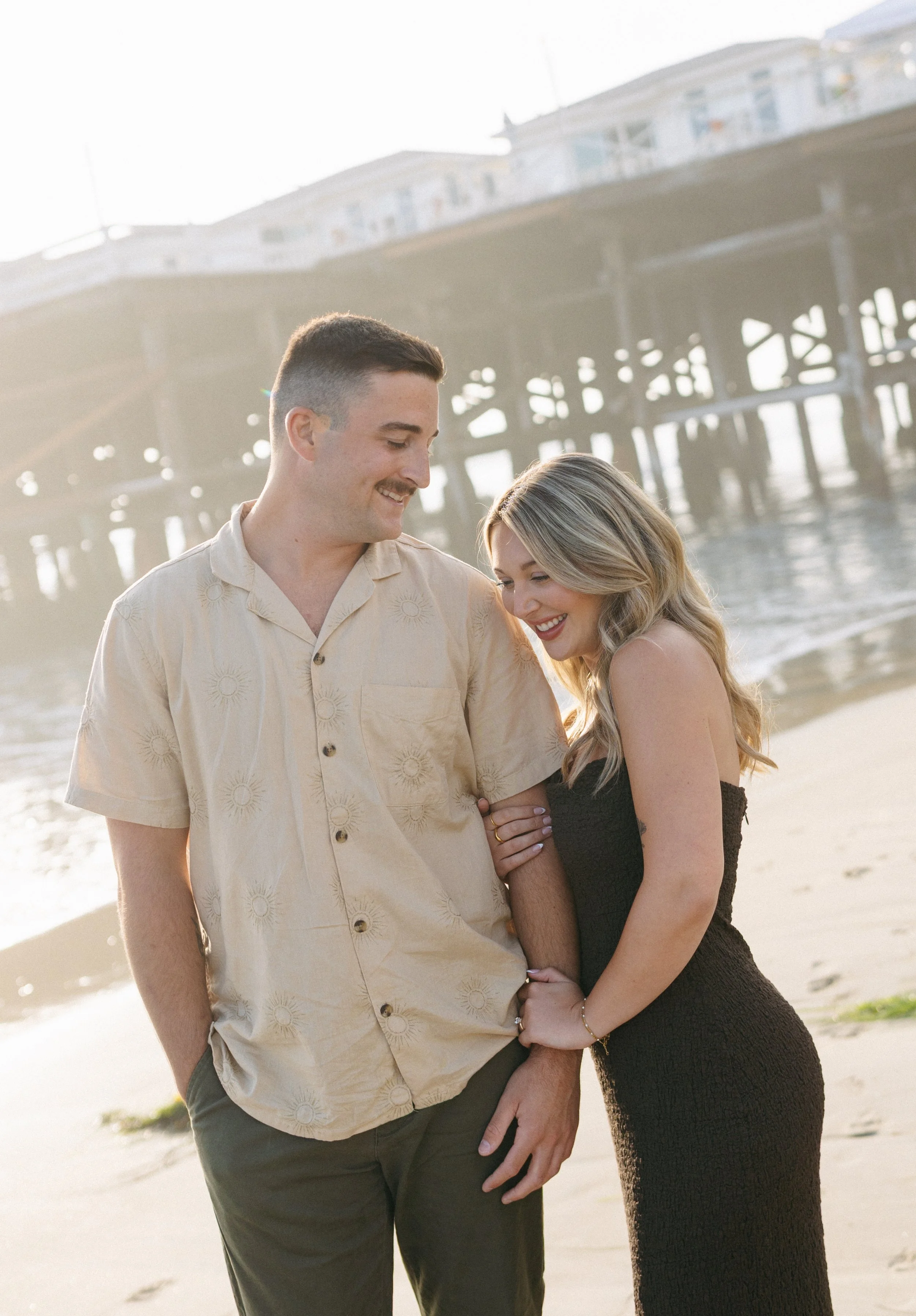 A happy couple stands on the beach near a pier, smiling and looking at each other, with sunlight shining behind them.