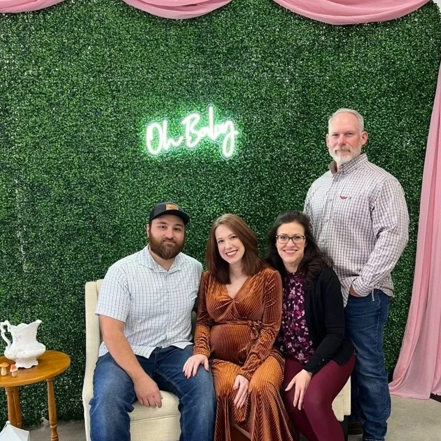 Expectant parents seated on ivory loveseat in front of a boxwood greenery wall
