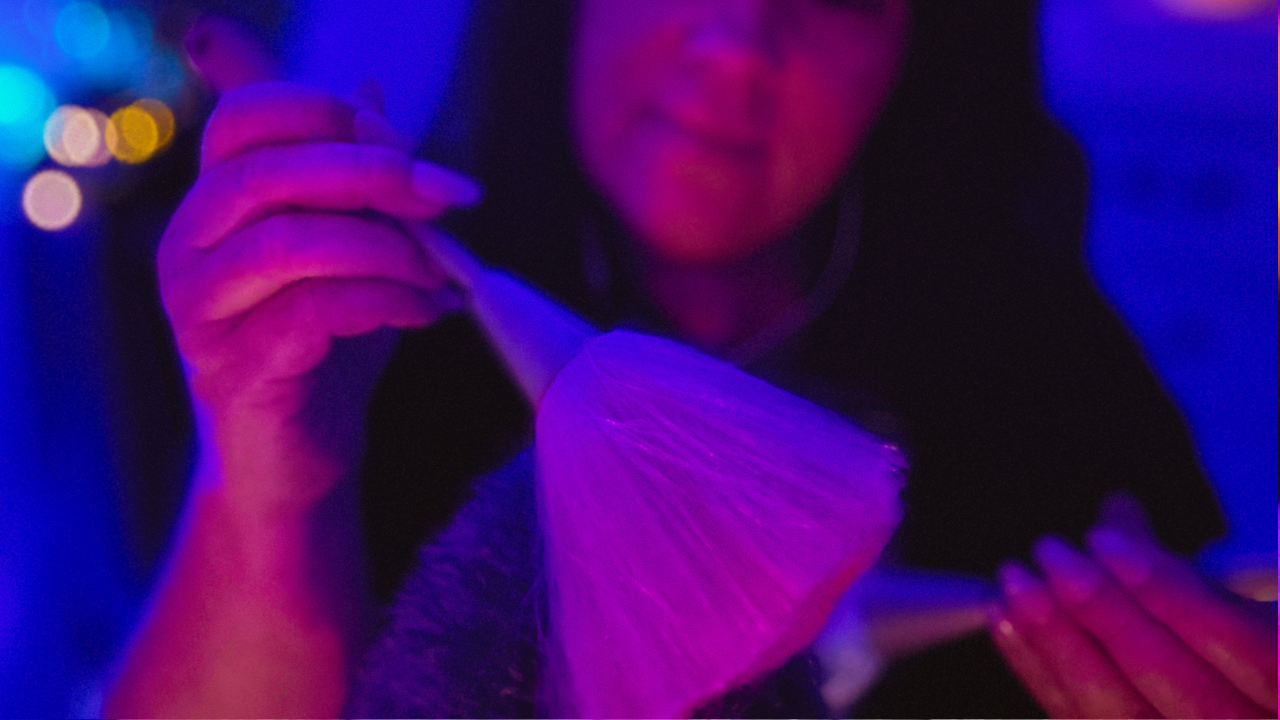 Woman gently brushing a microphone with a soft makeup brush in a calming purple-lit ASMR setting, creating relaxing mic brushing sounds for sleep, stress relief, and nervous system relaxation.
