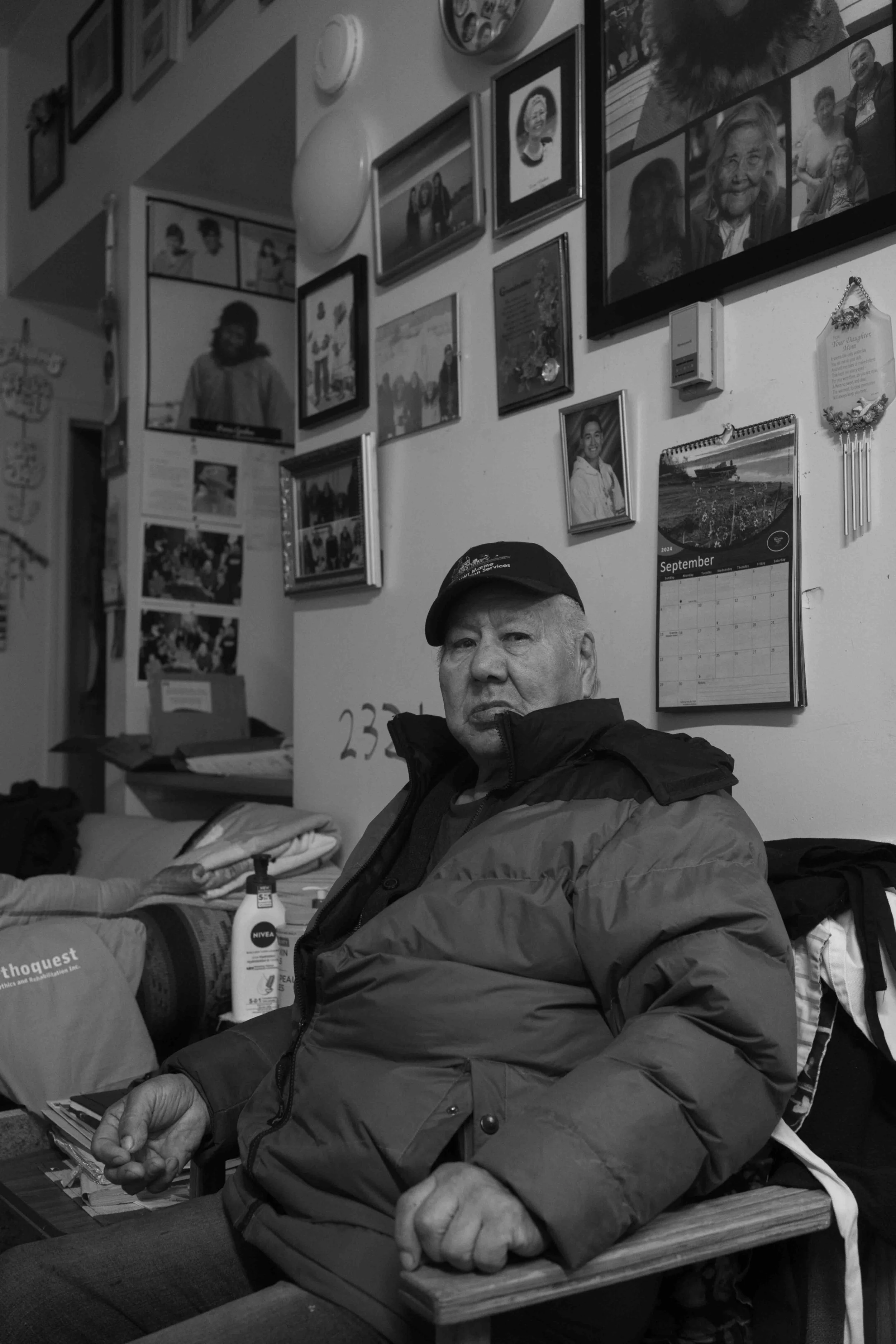 Black and white photo of an elderly man sitting in a room decorated with numerous framed photographs and pictures on the wall behind him.