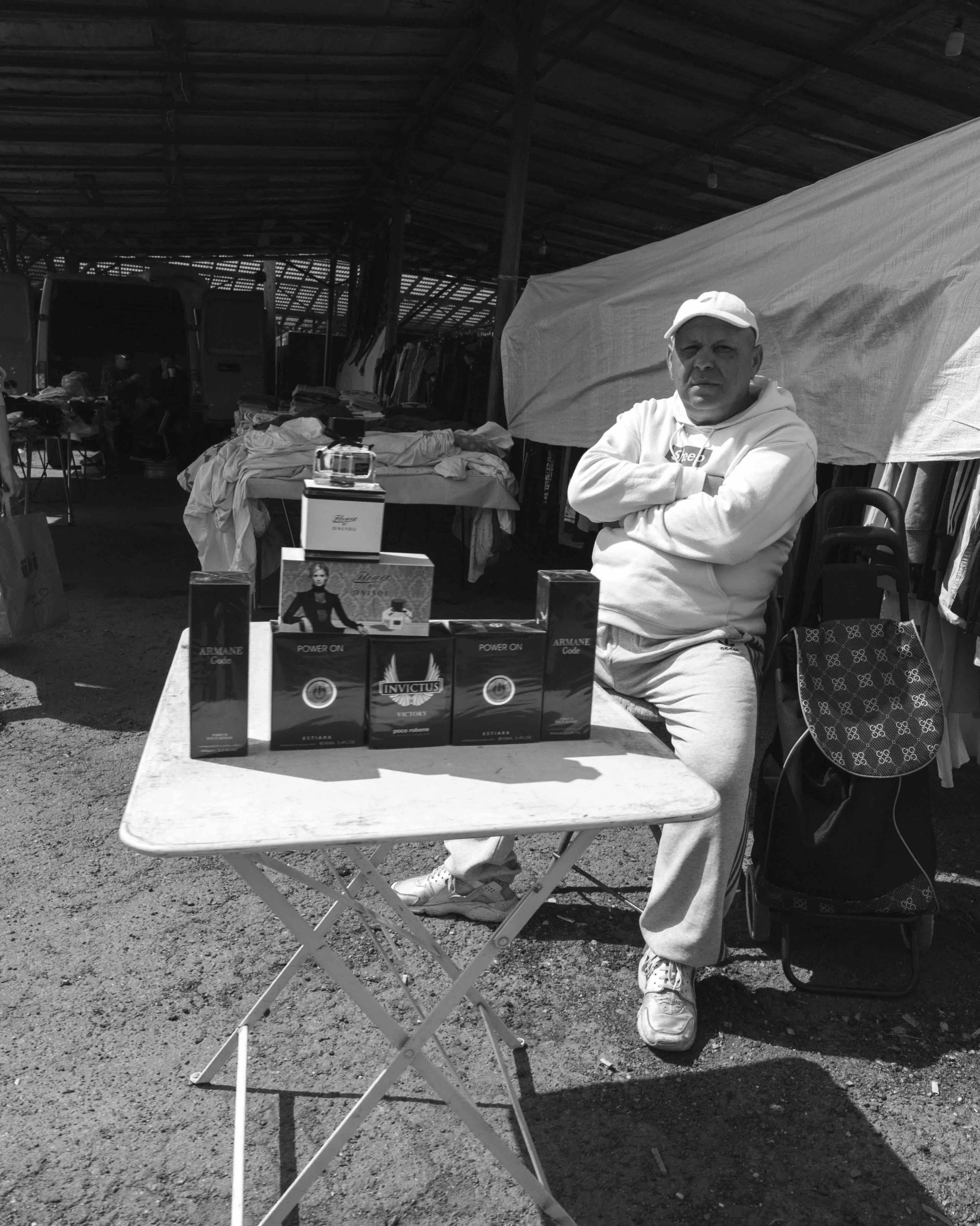 Man in white hoodie and light sweatpants standing behind a table with perfume bottles at an outdoor market.