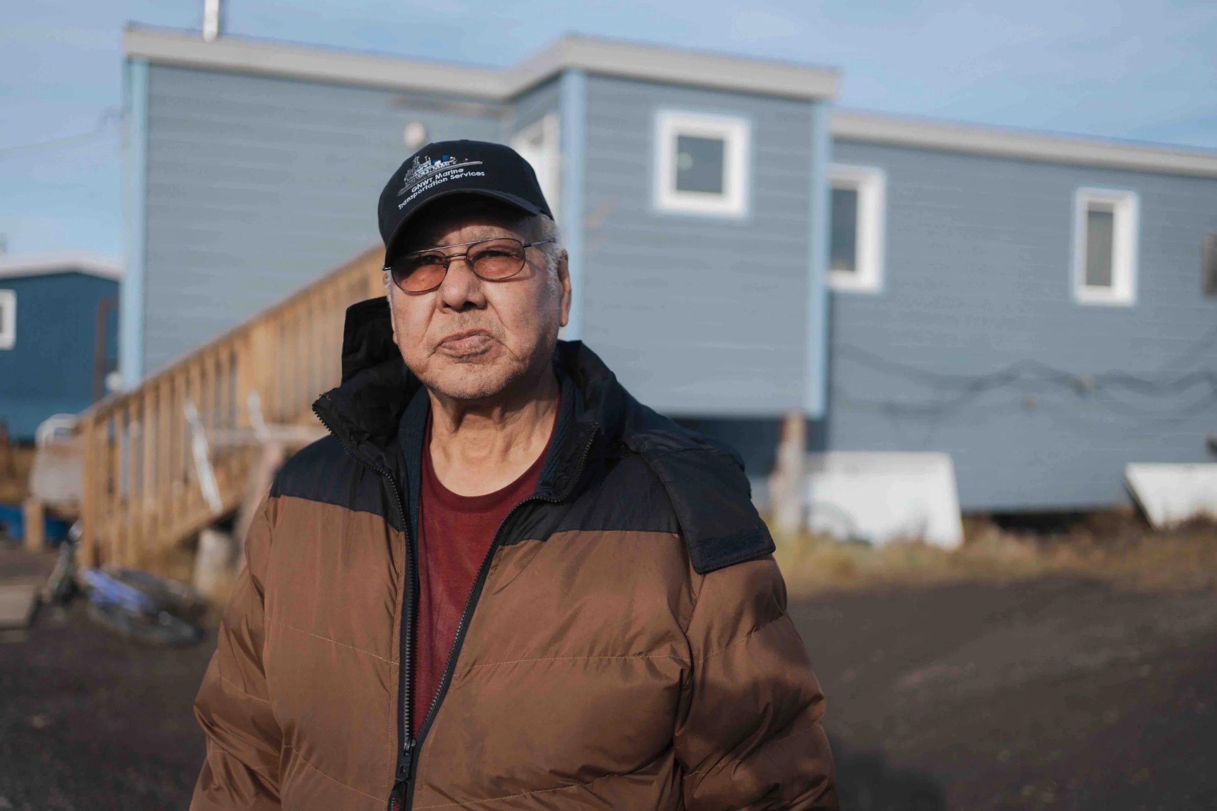 Older man in sunglasses and a black baseball cap with a brown and black jacket standing outdoors, with a modern blue house in the background.