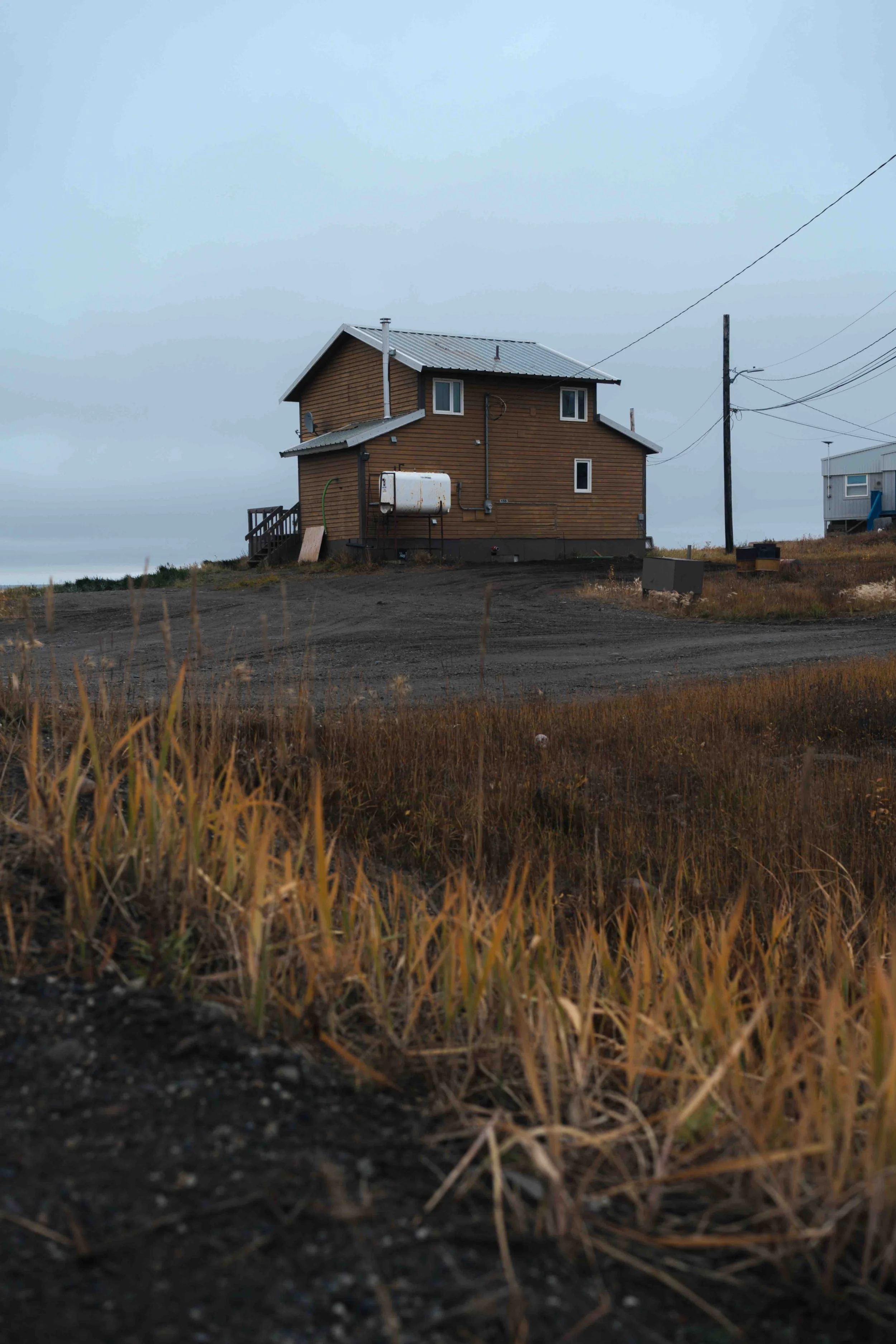 A house with a metal roof and wooden siding situated on a dirt lot with dry grass in the foreground, electrical lines overhead, and other houses visible in the distance under a gray sky.