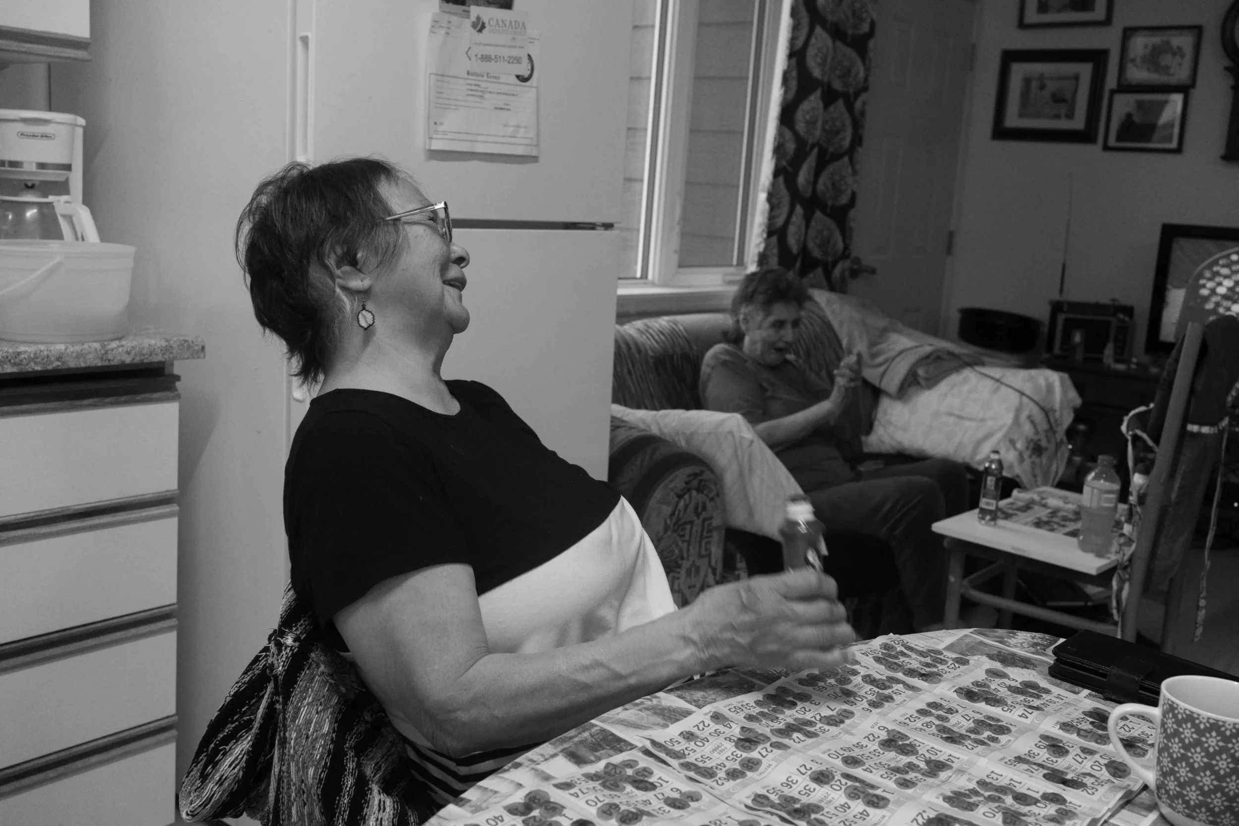 Two elderly women sitting in a living room, one at a dining table with lottery tickets, and the other on a couch, watching television, with drinks and snacks on the table.