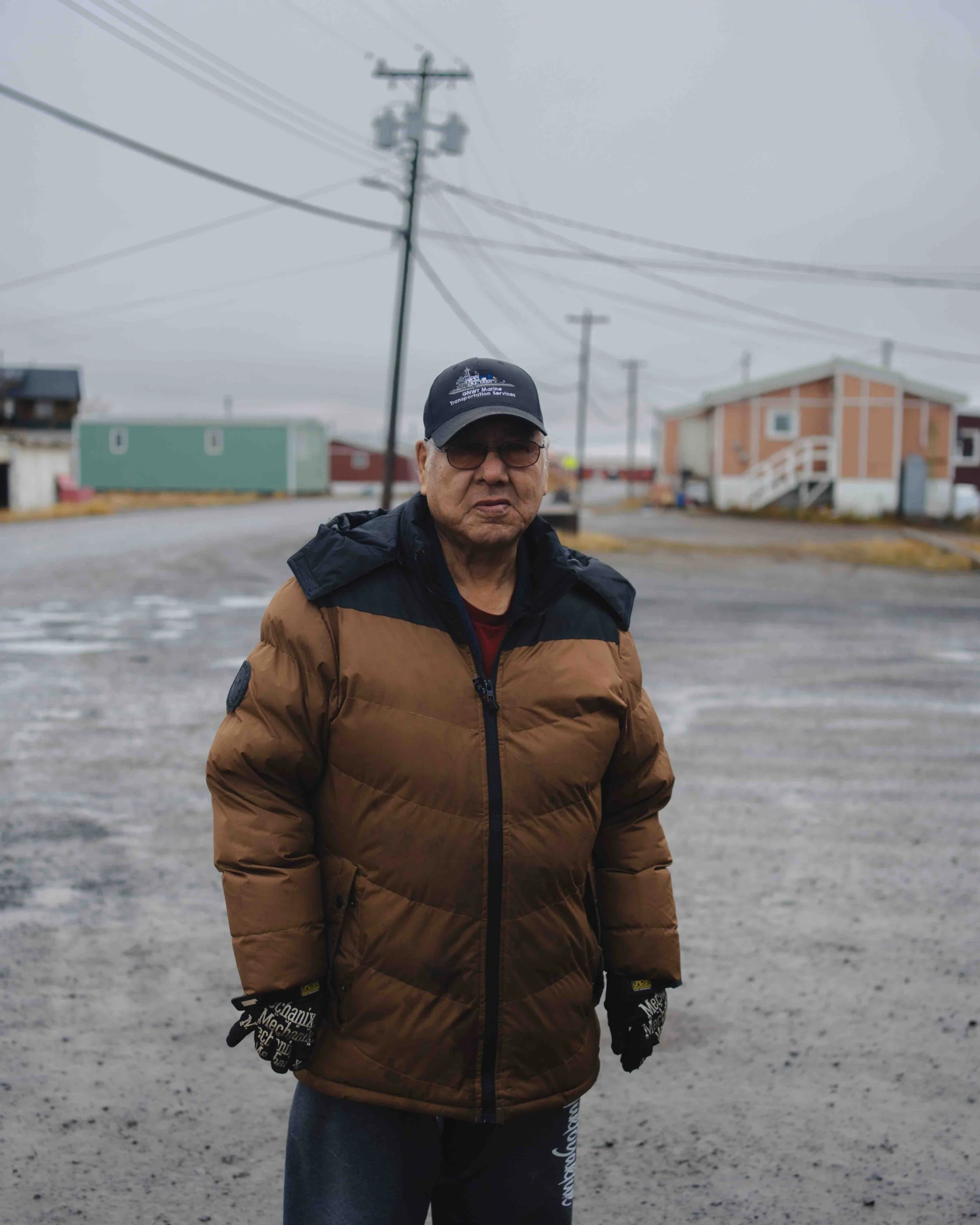 A man wearing a brown jacket, black gloves, glasses, and a baseball cap stands on a wet, gray street with utility poles and small houses in the background under cloudy skies.