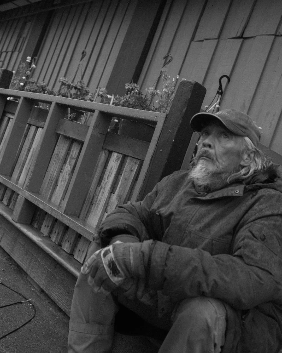 A black and white photo of an older man with a beard and long hair, wearing a baseball cap and jacket, sitting on the ground with his arm resting on his knee in a rustic setting with wooden panels and flower boxes.