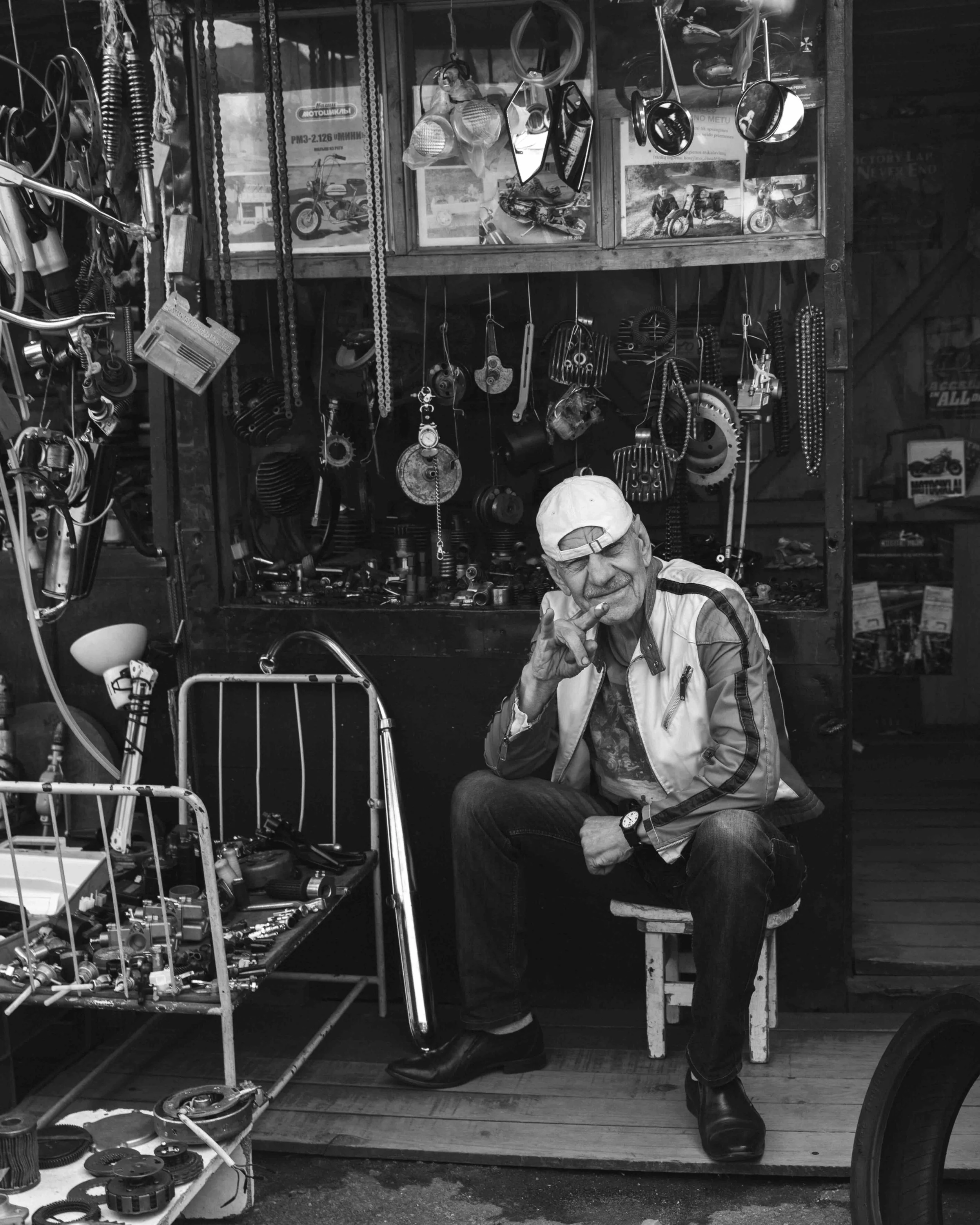 Black and white photo of an elderly man sitting in a cluttered workshop with various tools and motorcycle parts hanging on the wall behind him, making a peace sign gesture with his right hand.