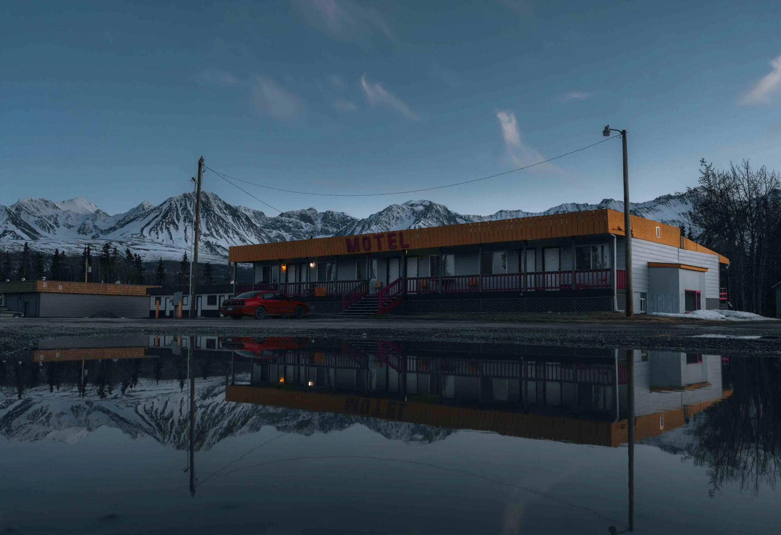 A motel building with a sign that reads 'MOTEL' in purple letters, a red car parked in front, and snow-capped mountains in the background. The reflection of the motel and mountains is visible in a puddle in the foreground.