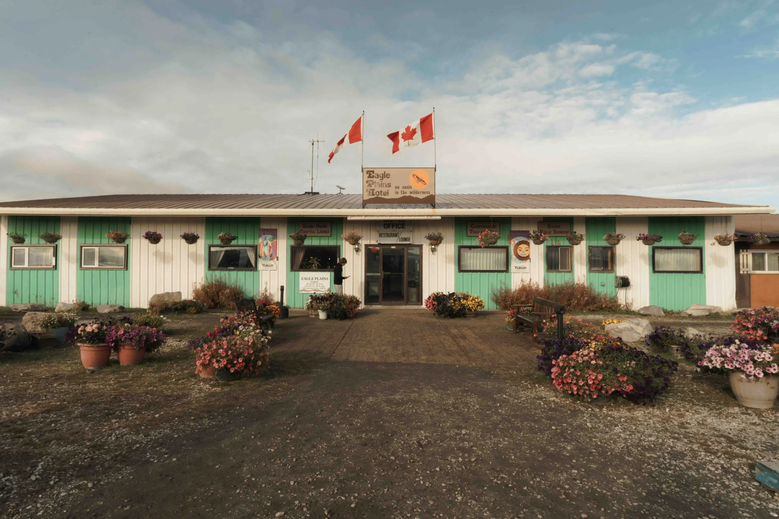 A single-story building with mint green and white exterior, decorated with flower pots and hanging flower baskets. Flags of Canada and the United States are flying on the roof. The building serves as an inn or lodge with signs for a restaurant and lo