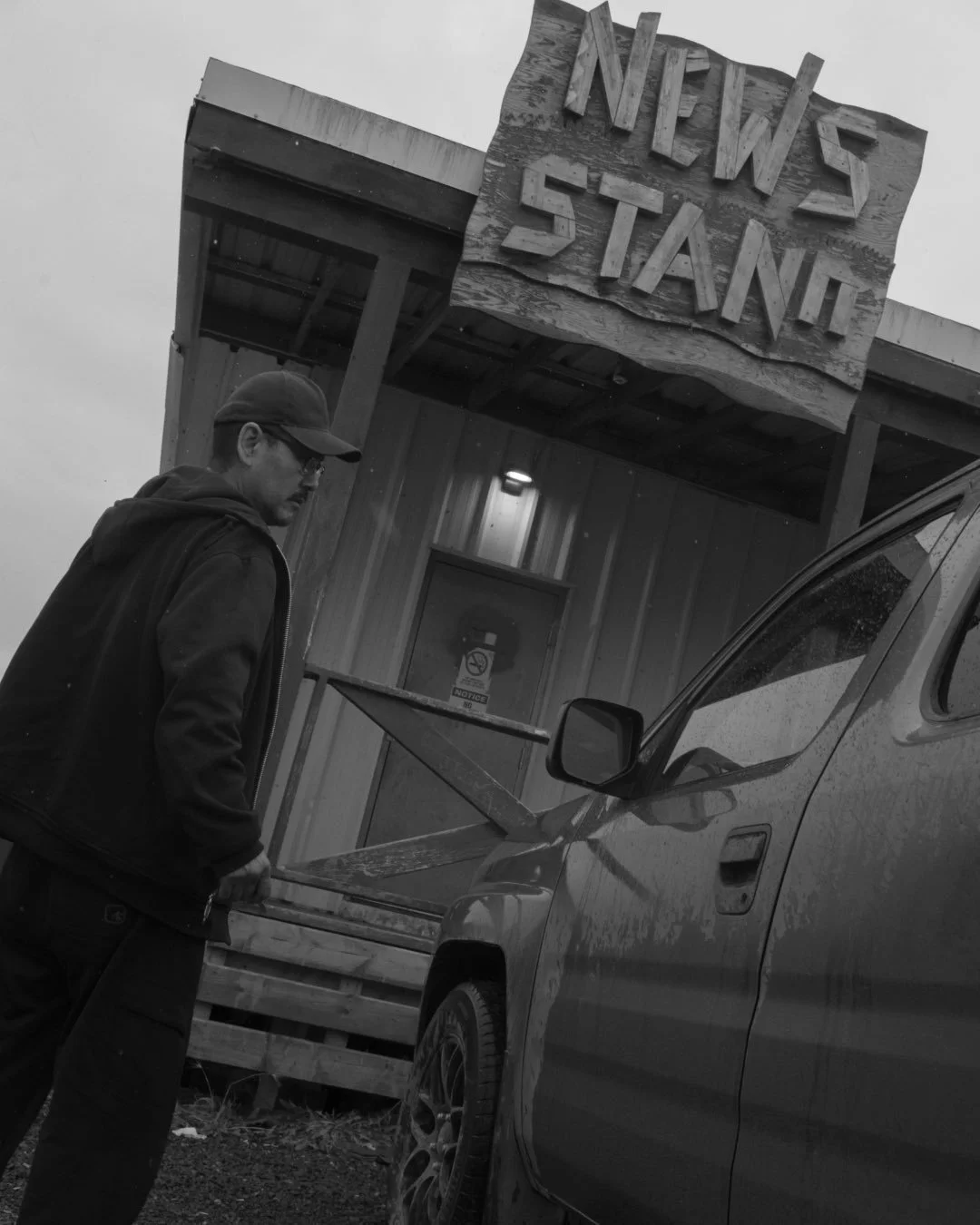 A man with a cap and jacket loading a tray into a vehicle in front of a building with a wooden sign that reads 'NEWS STAND'.