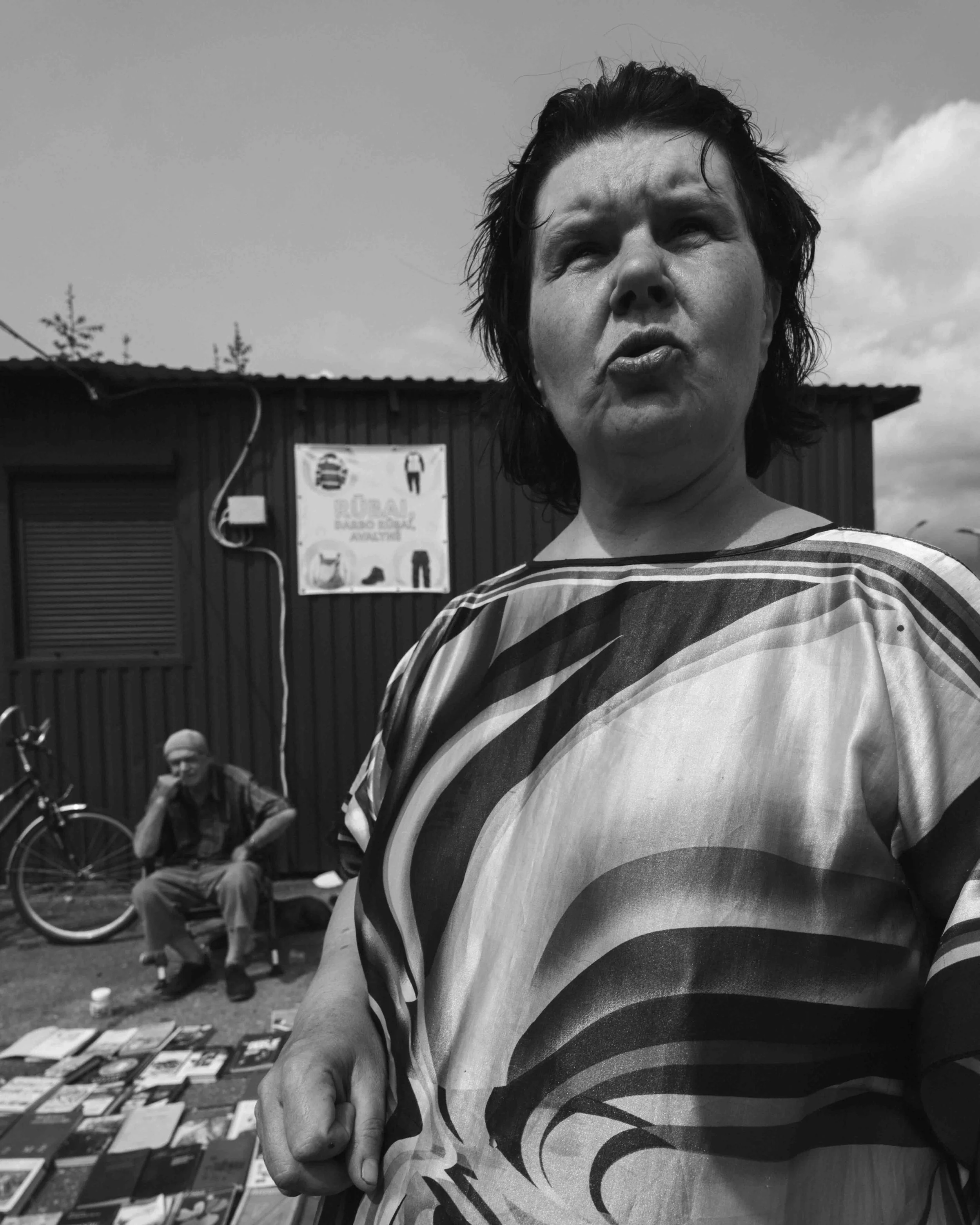 A woman in a swirling patterned top taking a photo outdoors with a man sitting on a bench and a bicycle in the background.