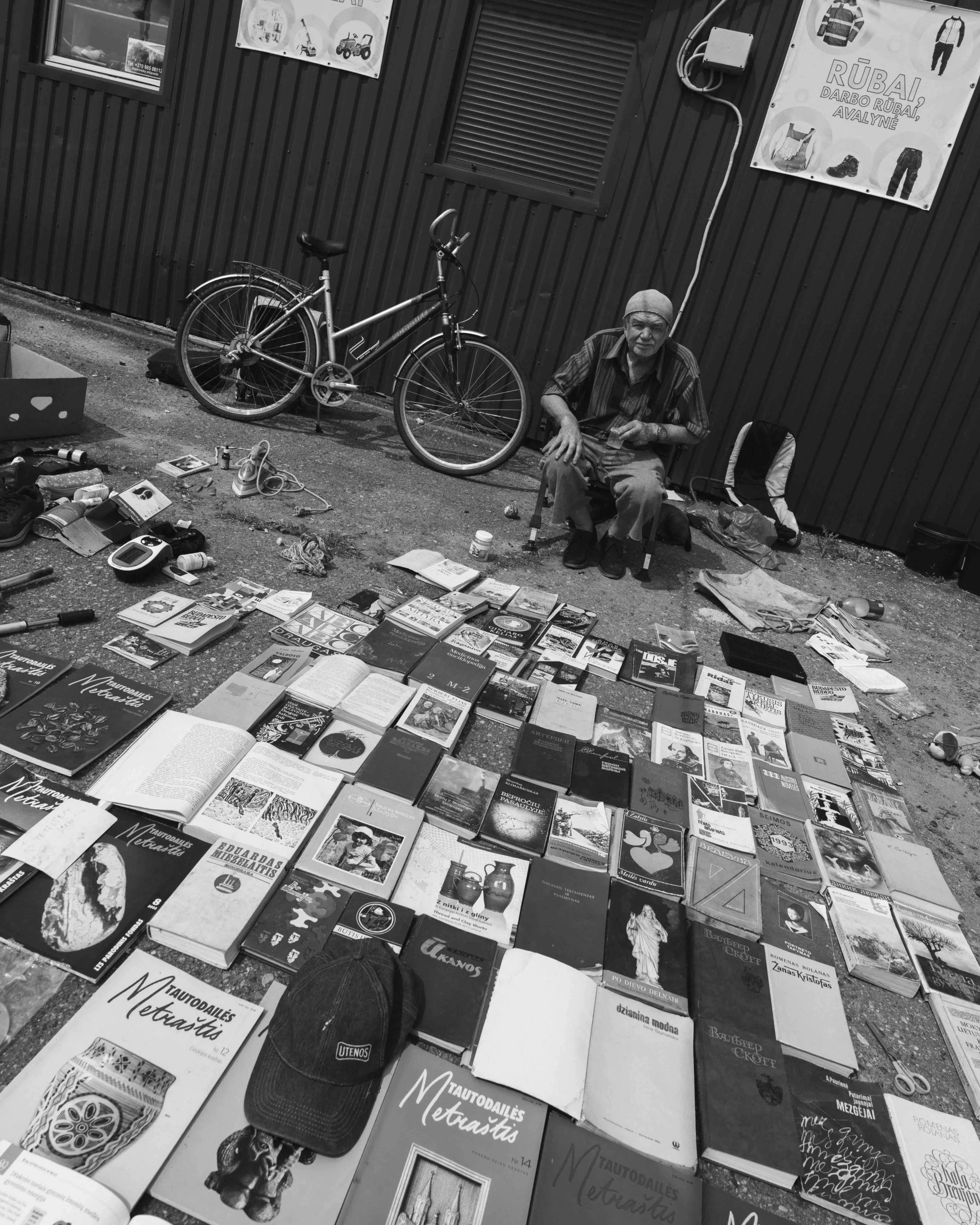 A man sitting on a small stool surrounded by books and items for sale on the ground, with a bicycle parked against a dark metal building in the background. The scene appears to be a street market.