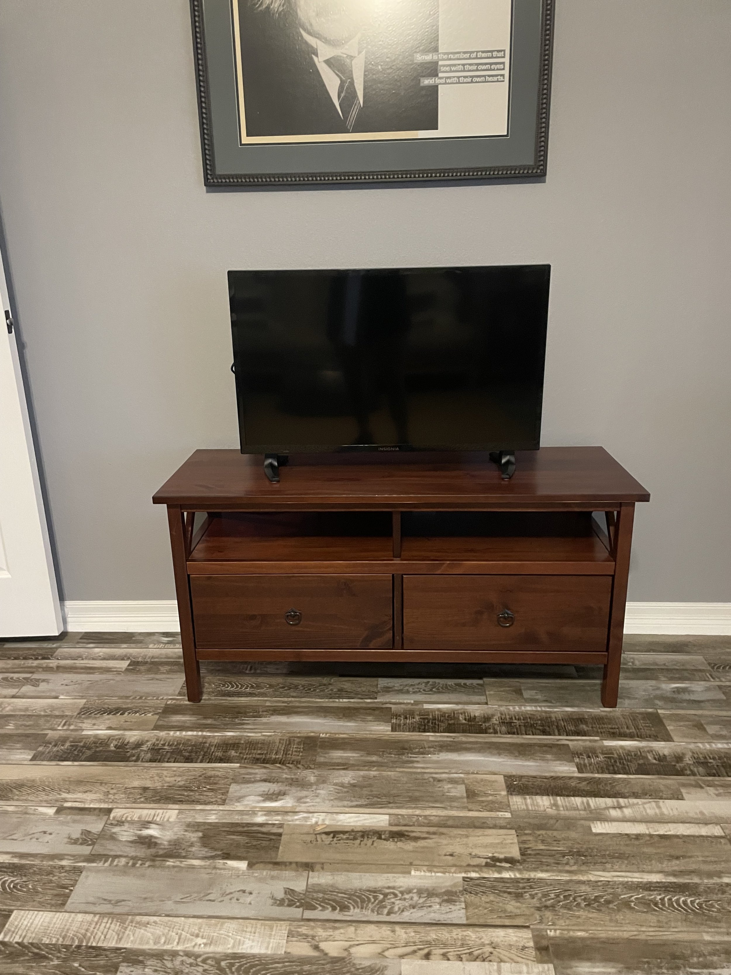 A flat-screen television on a wooden stand with two drawers and an open shelf, against a gray wall with a framed picture hanging above.