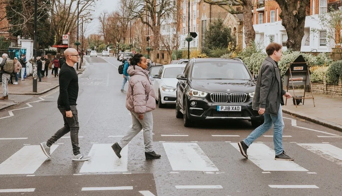 People crossing the street on a crosswalk in a city neighborhood with parked cars, trees, and residential buildings in the background.