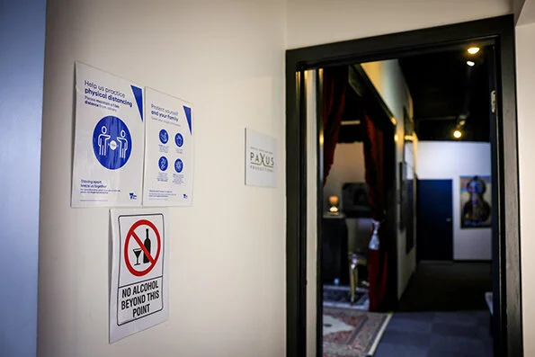 Interior hallway with signs about physical distancing, safety guidelines, and a no alcohol sign on the wall, leading to an art gallery or room with artwork.