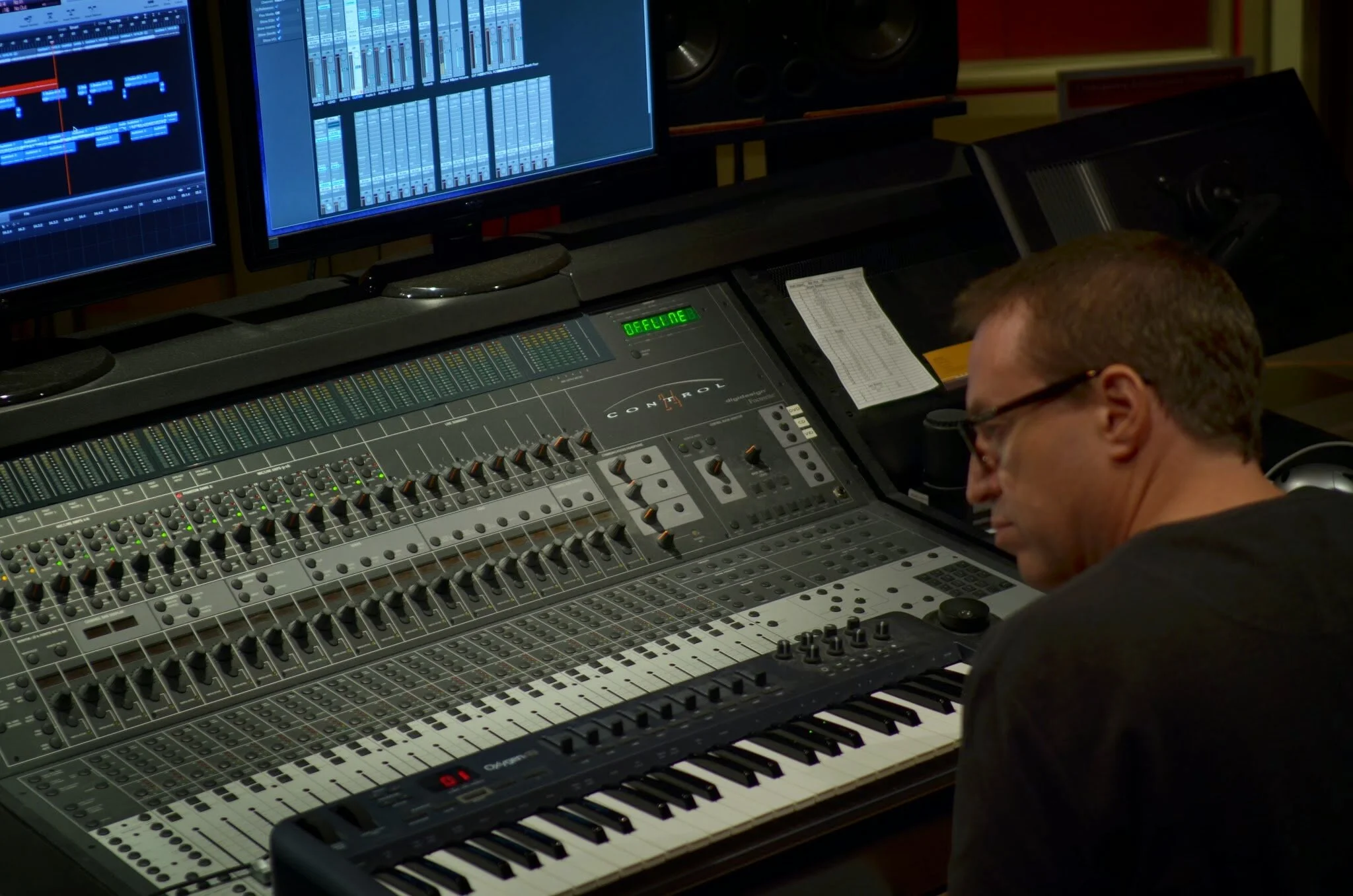 A man wearing glasses working with a large music mixing console with multiple knobs and sliders, in front of two monitors displaying digital audio tracks and controls.