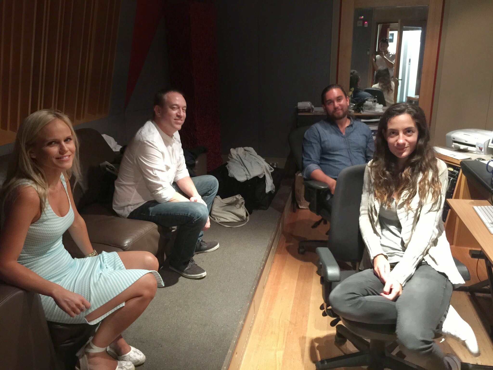 Four people in a recording studio, two women sitting on a couch and two men sitting on office chairs, with a woman taking a photo through a mirror